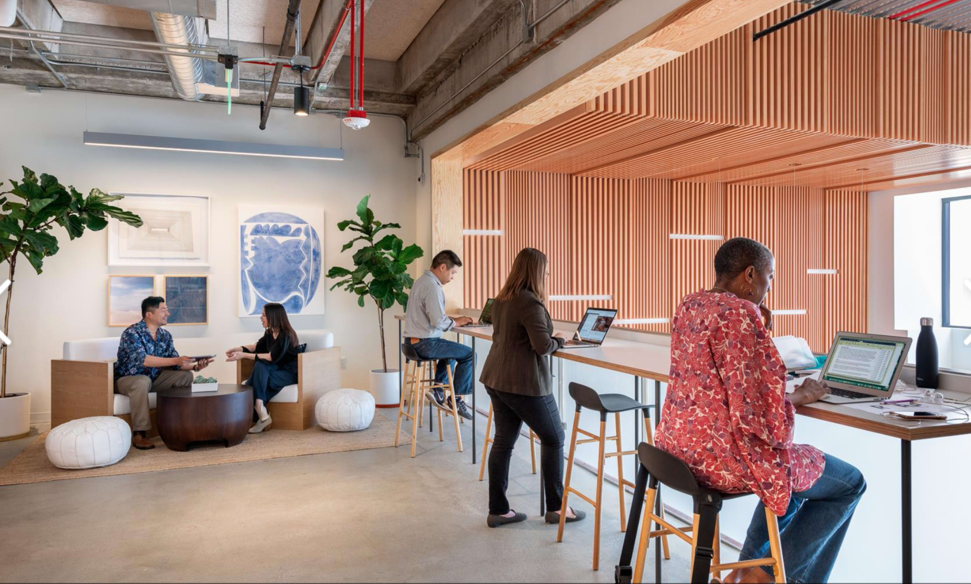 The image shows a modern office space with several people working on laptops at various seating areas, including a lounge with plants and wooden paneling.