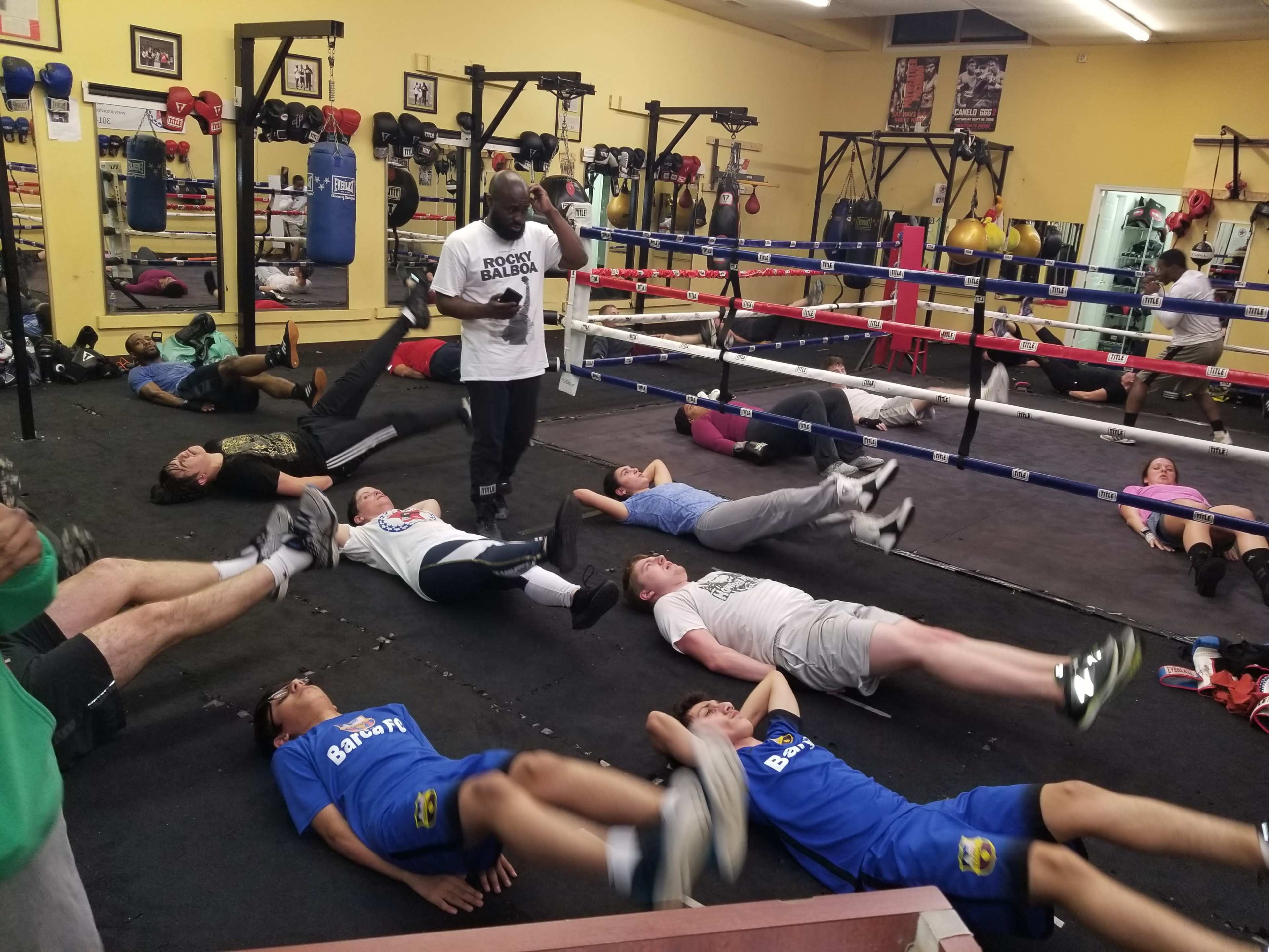 A group of people is participating in a boxing training session, with some lying on the mat performing leg raises while others are in the ring or near the boxing equipment.