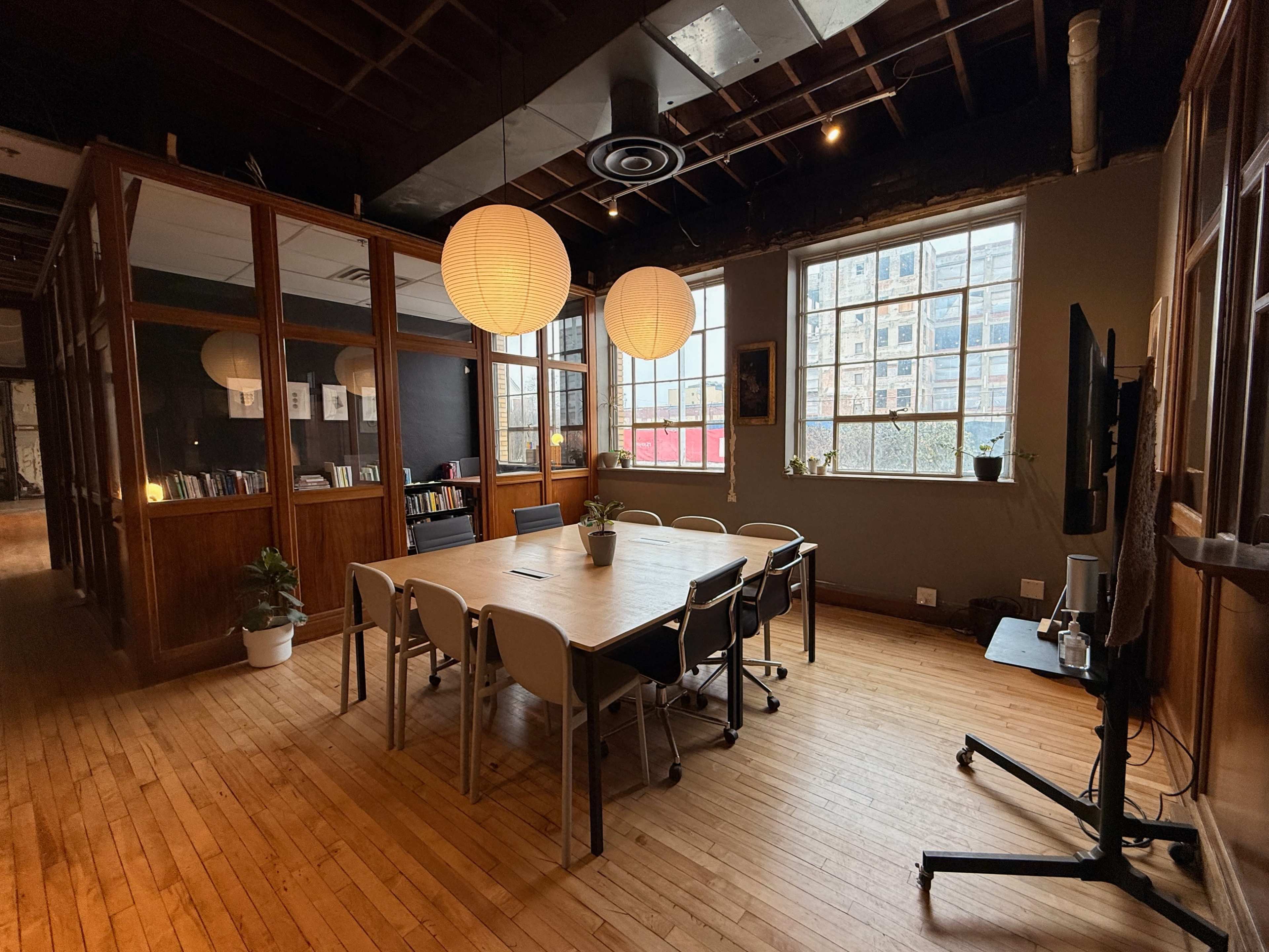The image shows a spacious meeting room with a large wooden table surrounded by chairs, illuminated by pendant lights and featuring large windows with a view of an urban environment.