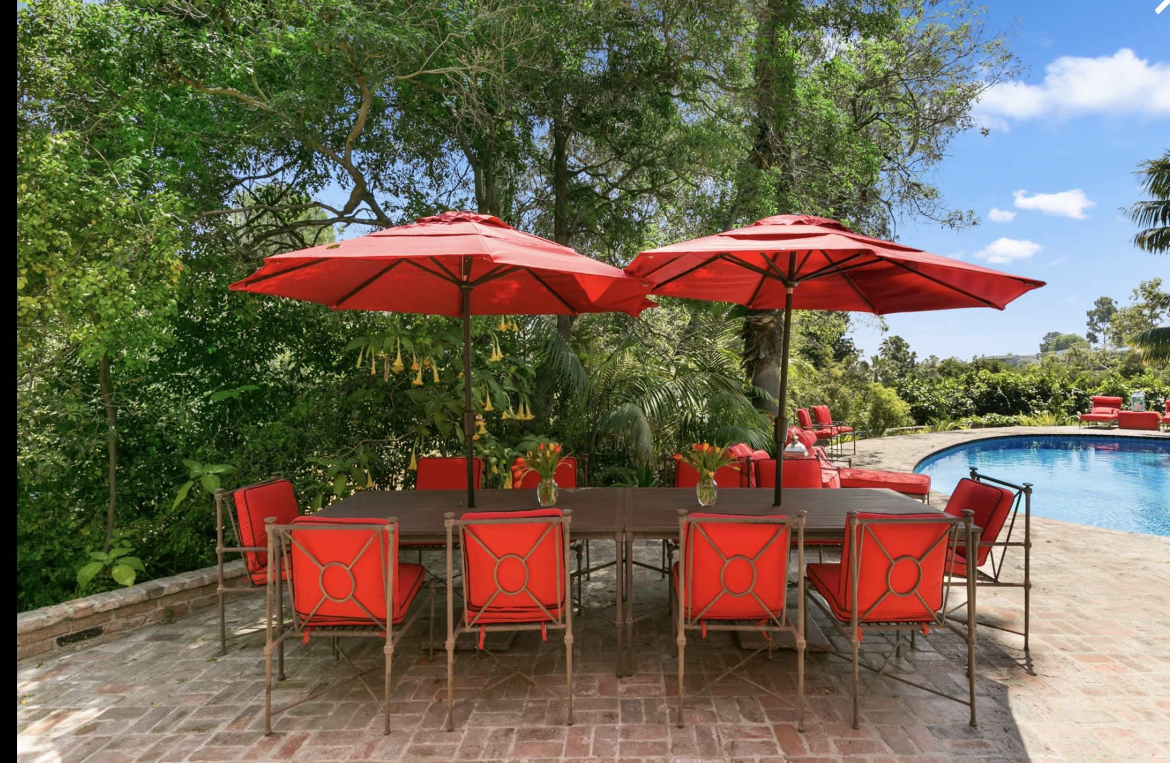 A dining table set with red chairs and umbrellas is placed near a swimming pool surrounded by lush greenery.