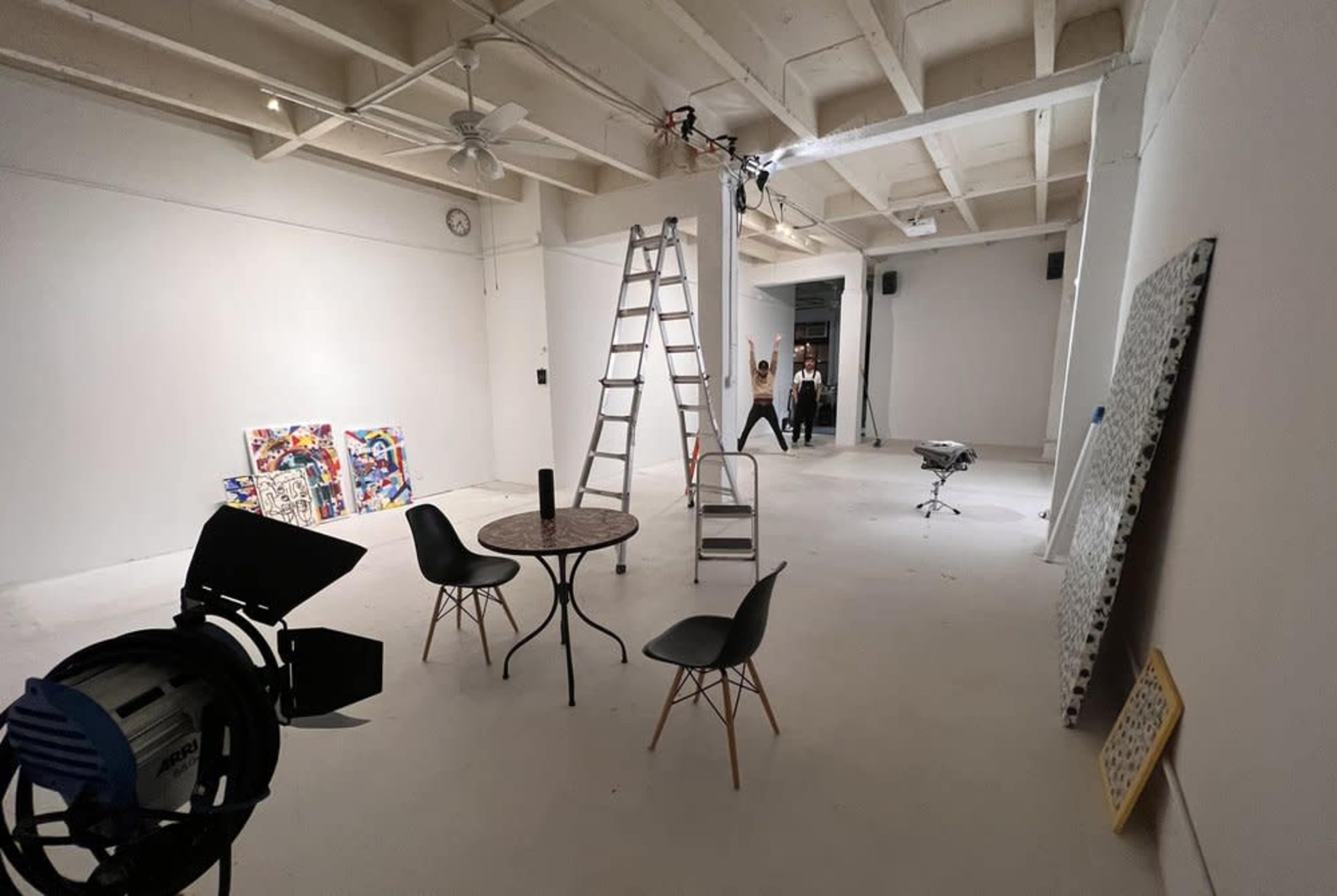 An artist's studio features a ladder, a table with chairs, an easel, and various art supplies in a spacious, well-lit room.
