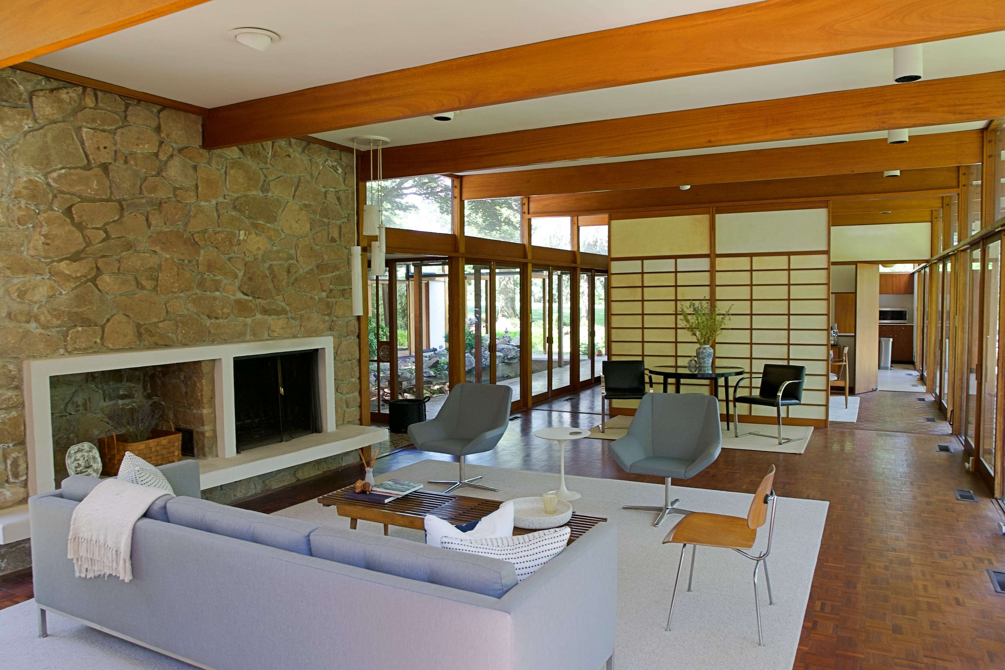 The image shows a modern living room with a stone fireplace, large windows, and minimalist furniture arranged around a central area rug.