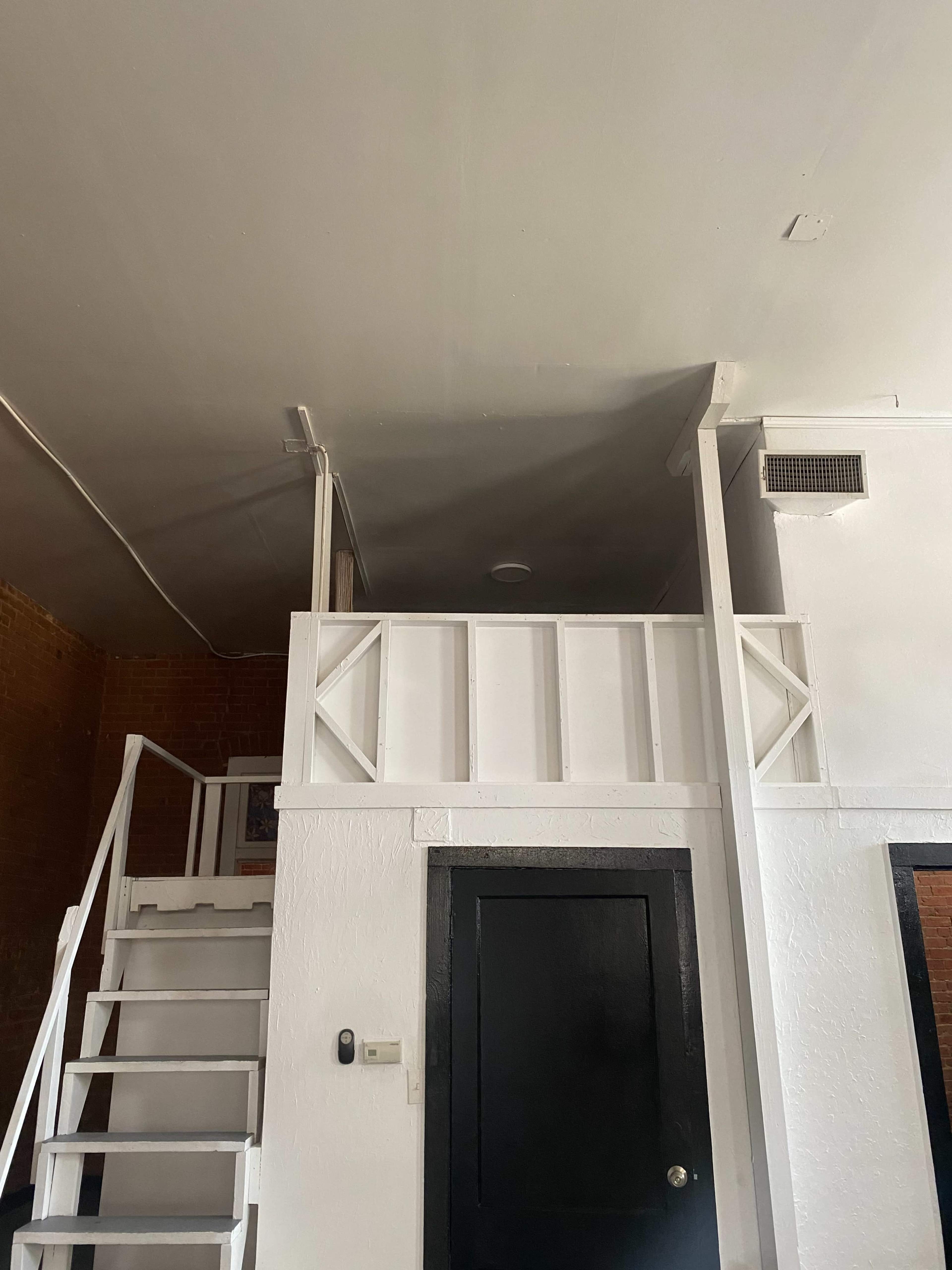 A staircase leading to a small loft area with a railing, alongside a black door on the wall.