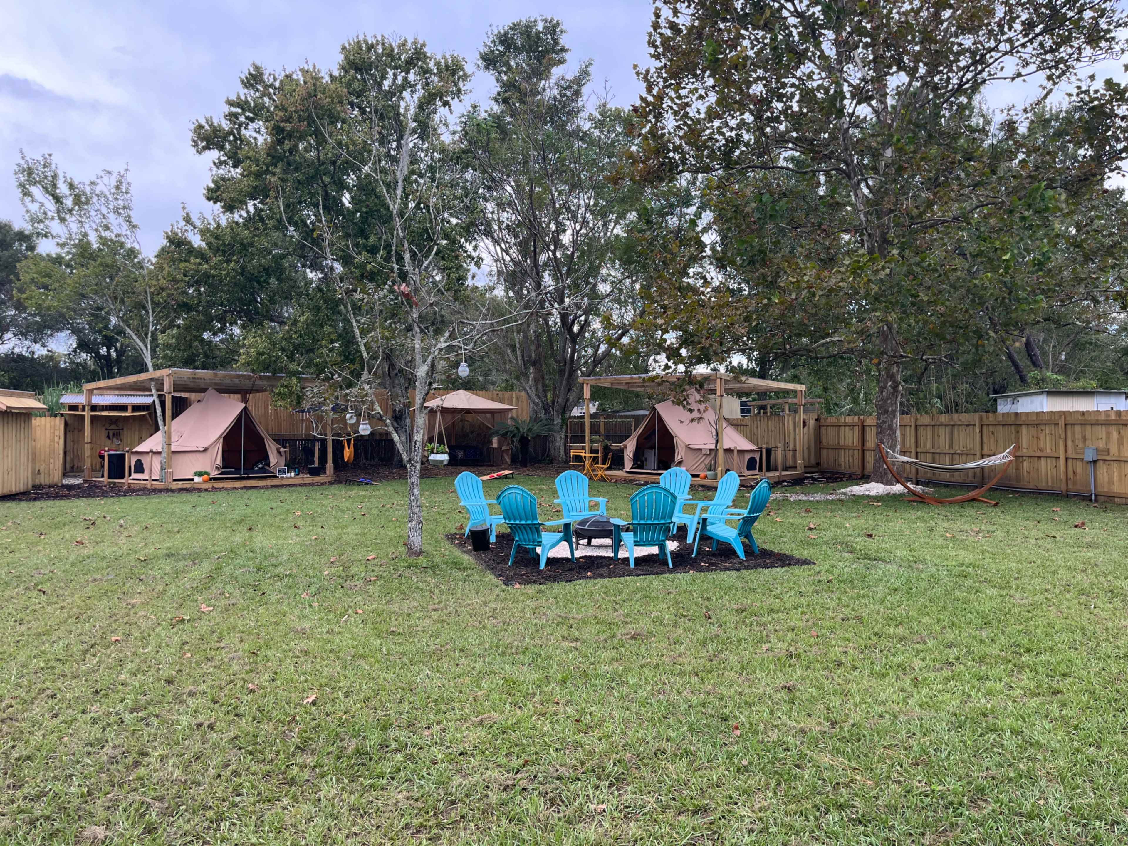 A grassy area with blue Adirondack chairs arranged around a fire pit, flanked by two tent-like structures and a hammock in a fenced yard.