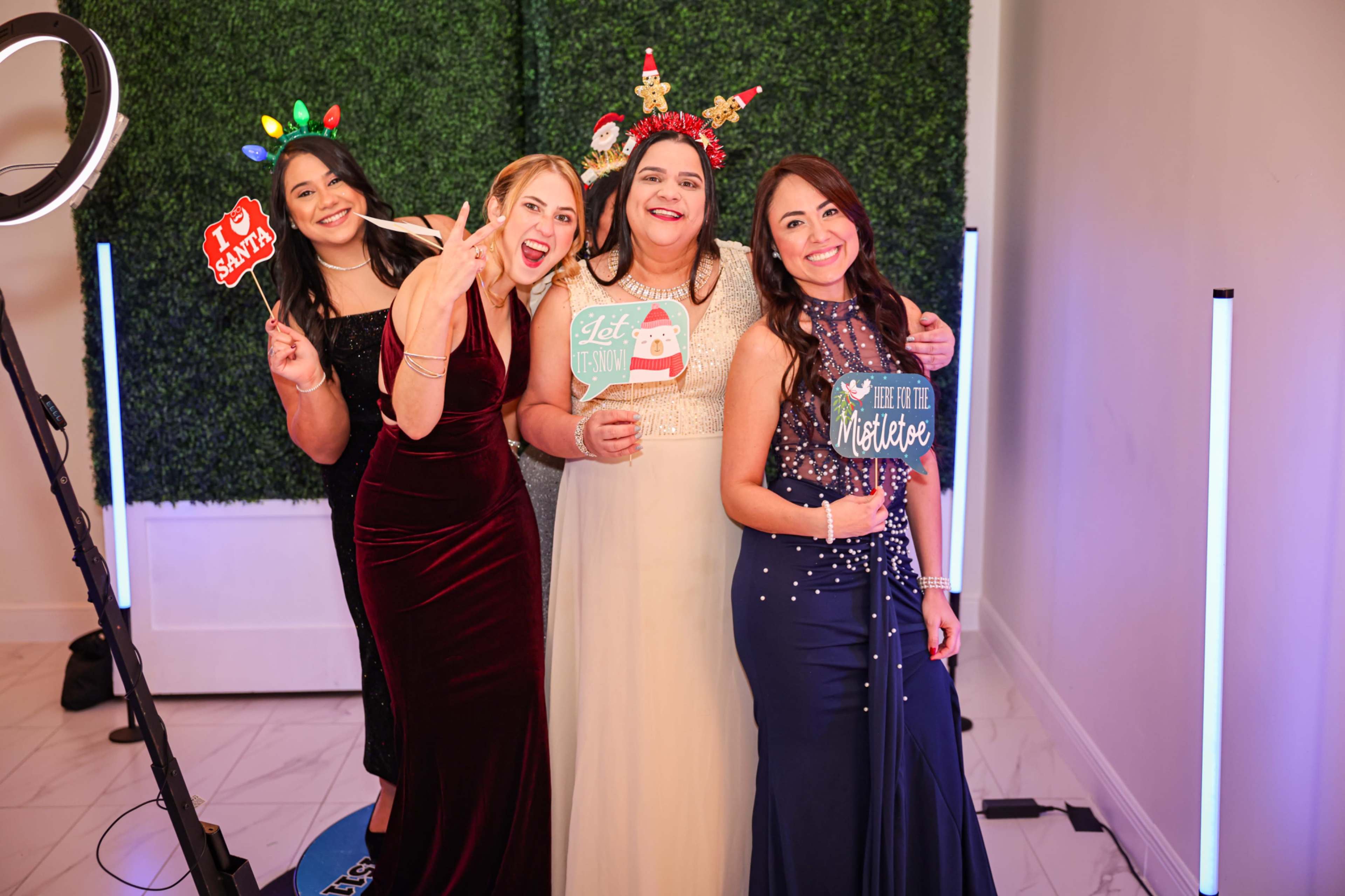 Four women pose together for a photo in front of a decorative green backdrop, each holding fun props and wearing festive attire.