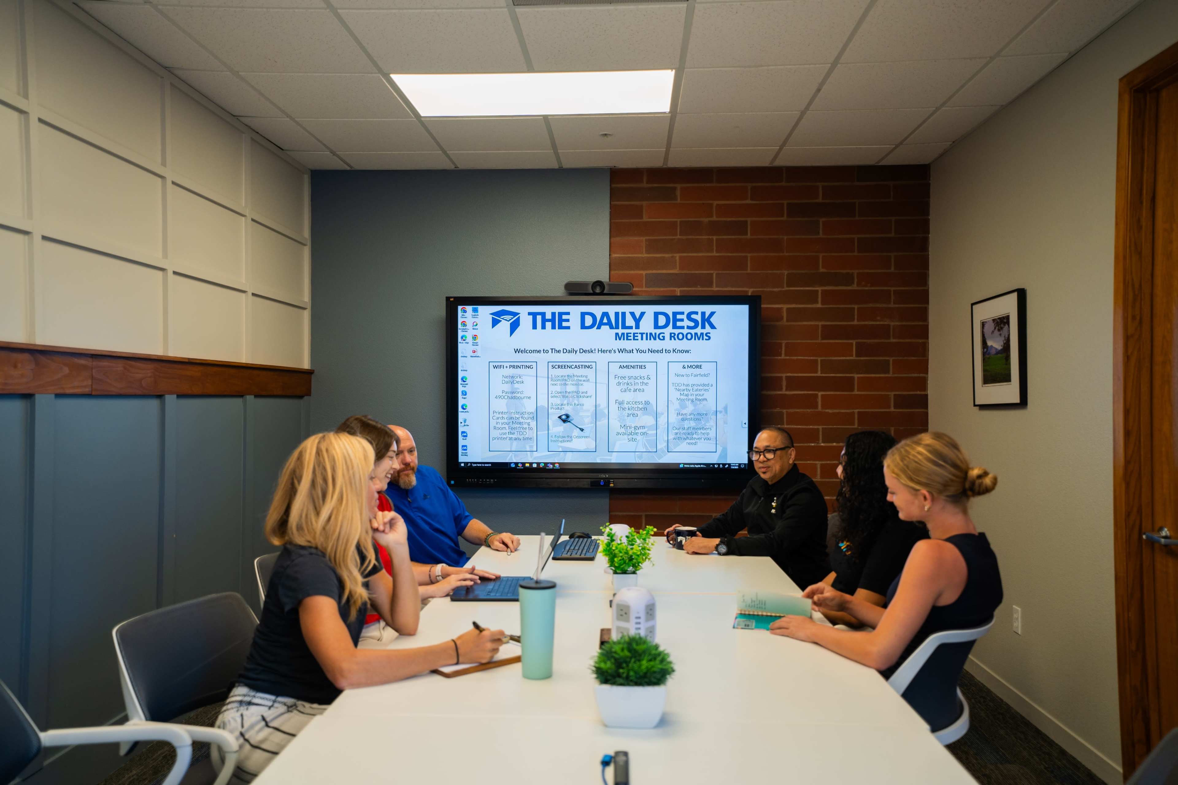 A group of six individuals sits around a conference table in a meeting room, with a large screen displaying "The Daily Desk Meeting Rooms" in the background.