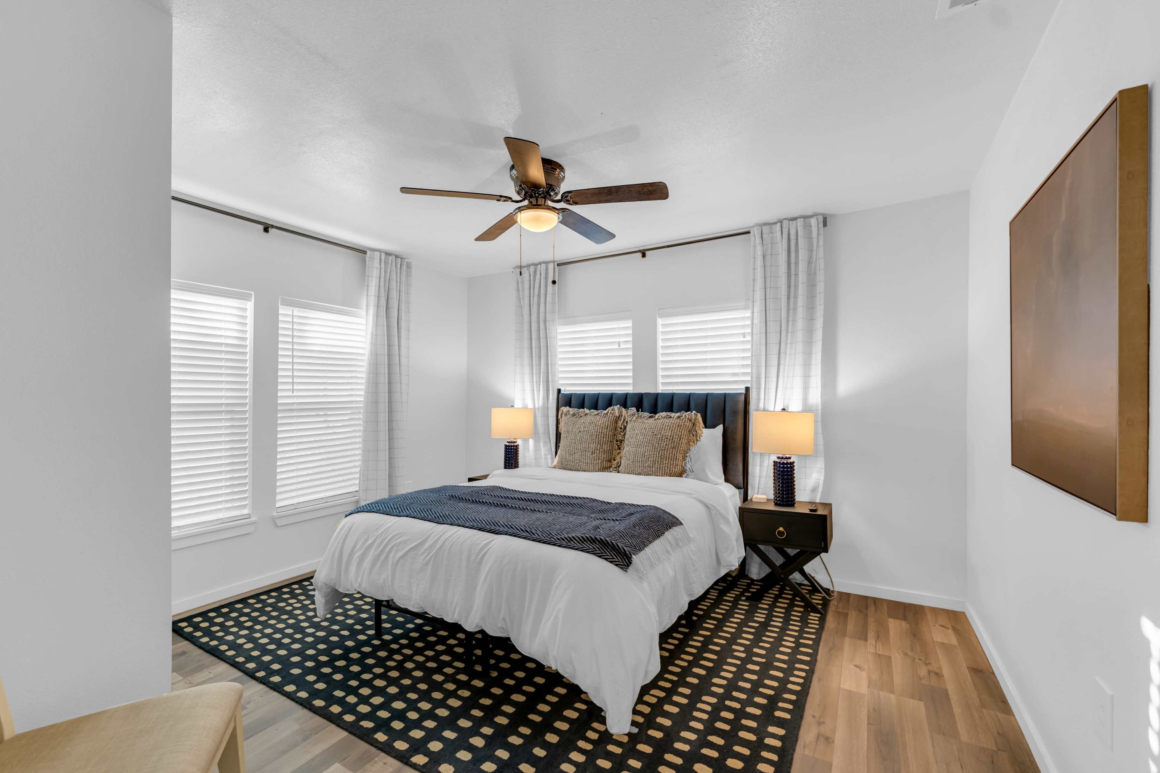 A well-lit bedroom featuring a bed with a white comforter, two lamps on nightstands, and large windows with white blinds.