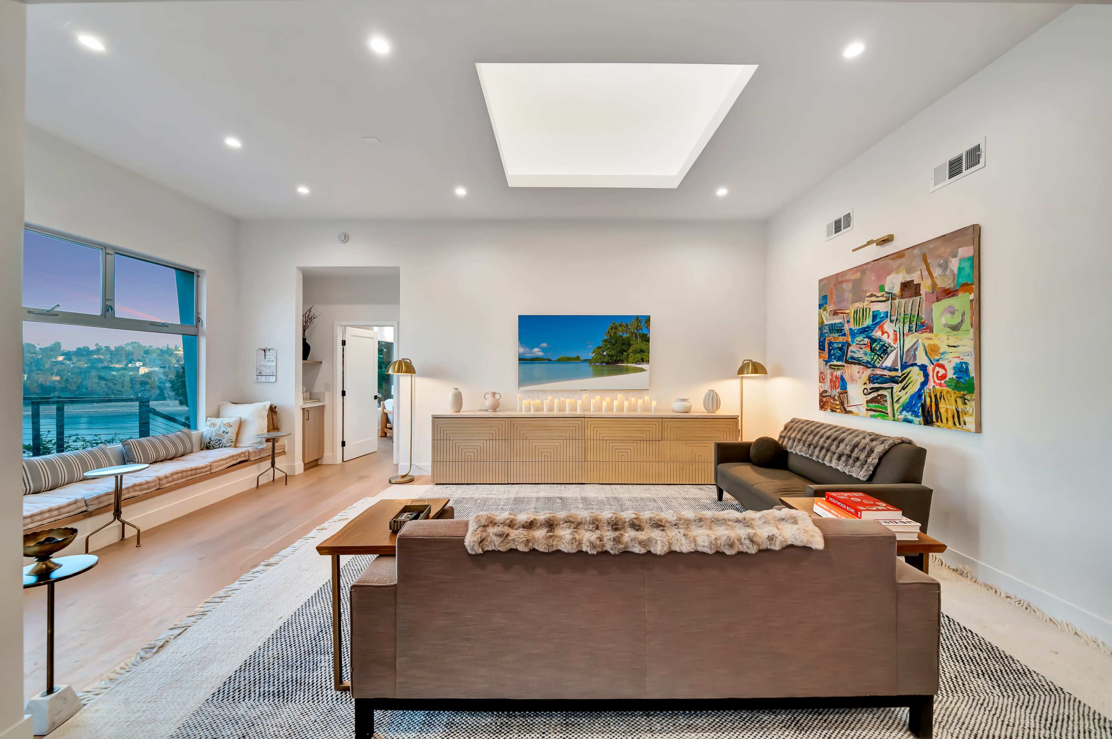The image shows a modern living room with a light-colored sofa, a dark coffee table, and an illuminated cabinet, featuring large windows that overlook a water view.
