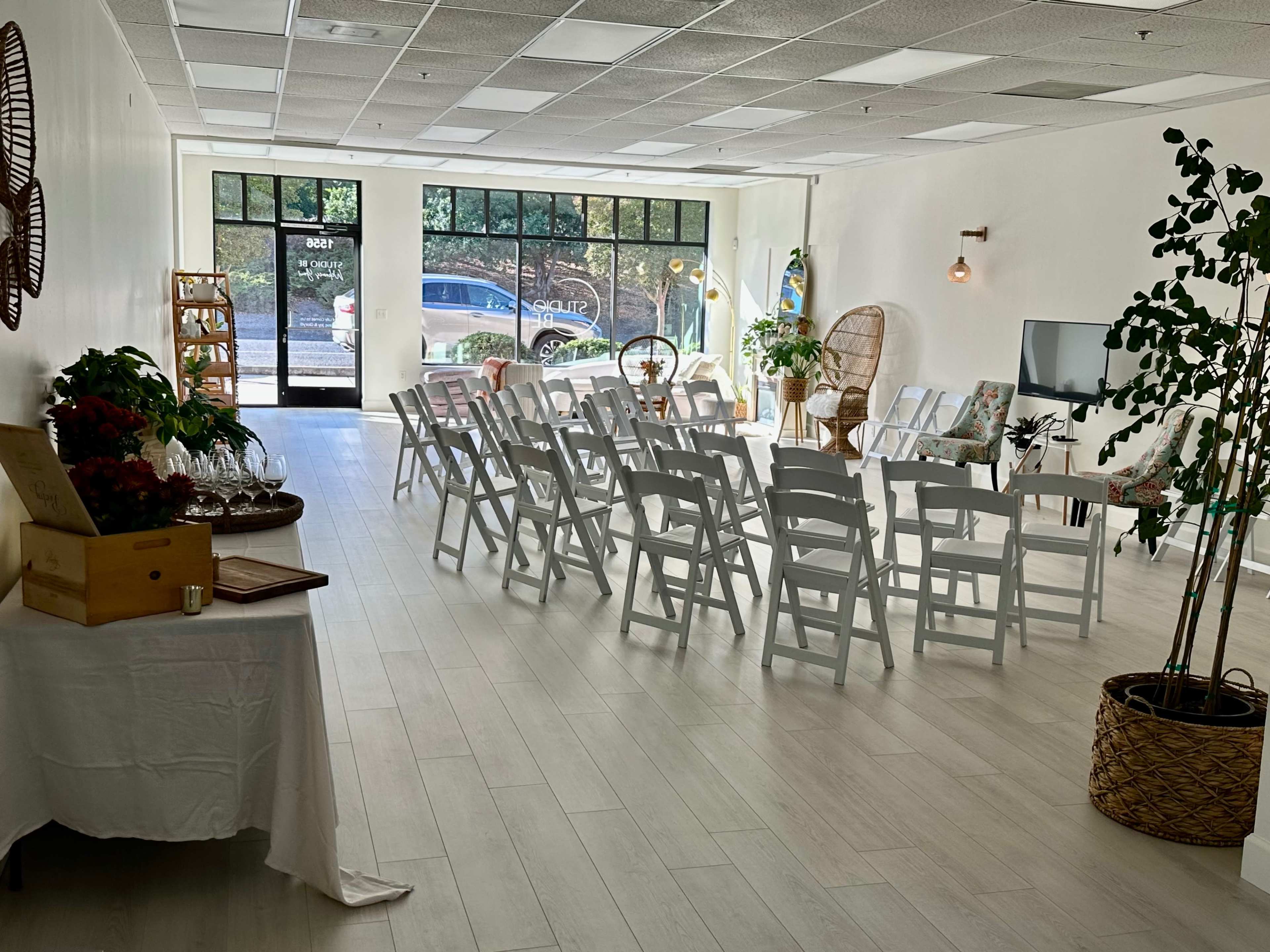 A light-filled event space set up with white folding chairs facing a small stage, surrounded by decorative plants and a side table with refreshments.