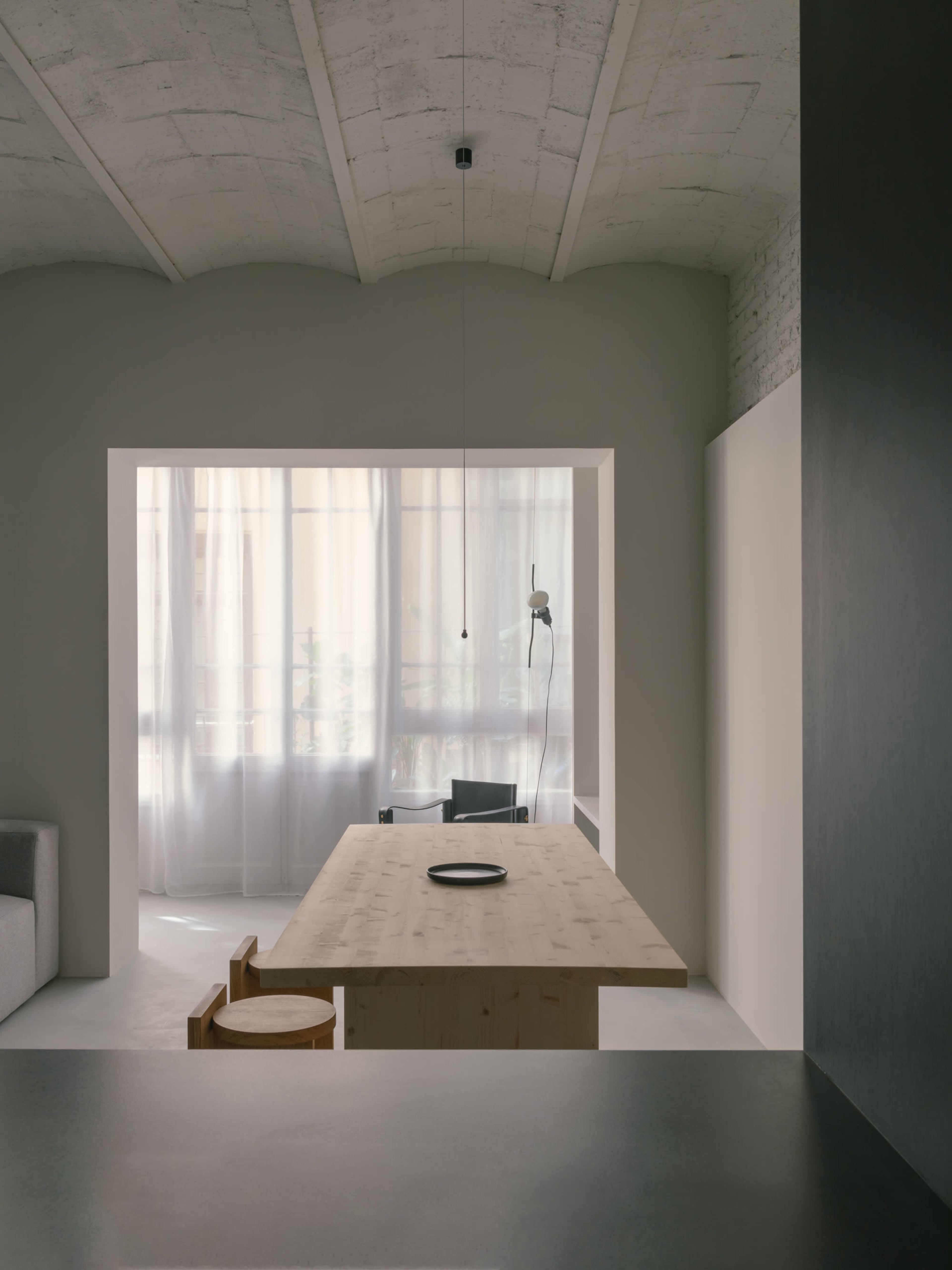 A minimalist dining area features a wooden table with a single plate and chairs, framed by sheer curtains and a softly lit background.