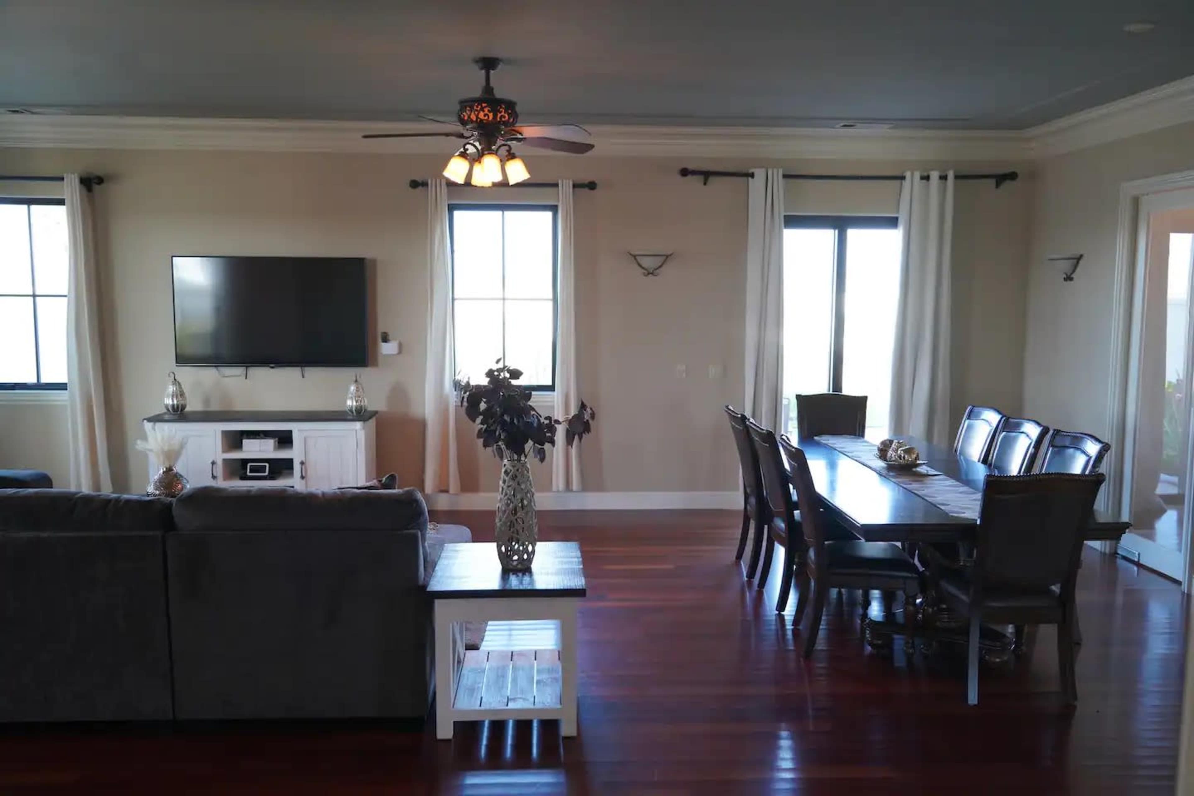 A spacious living and dining area featuring a large gray sofa, a wooden dining table with chairs, a television mounted on the wall, and large windows with curtains.