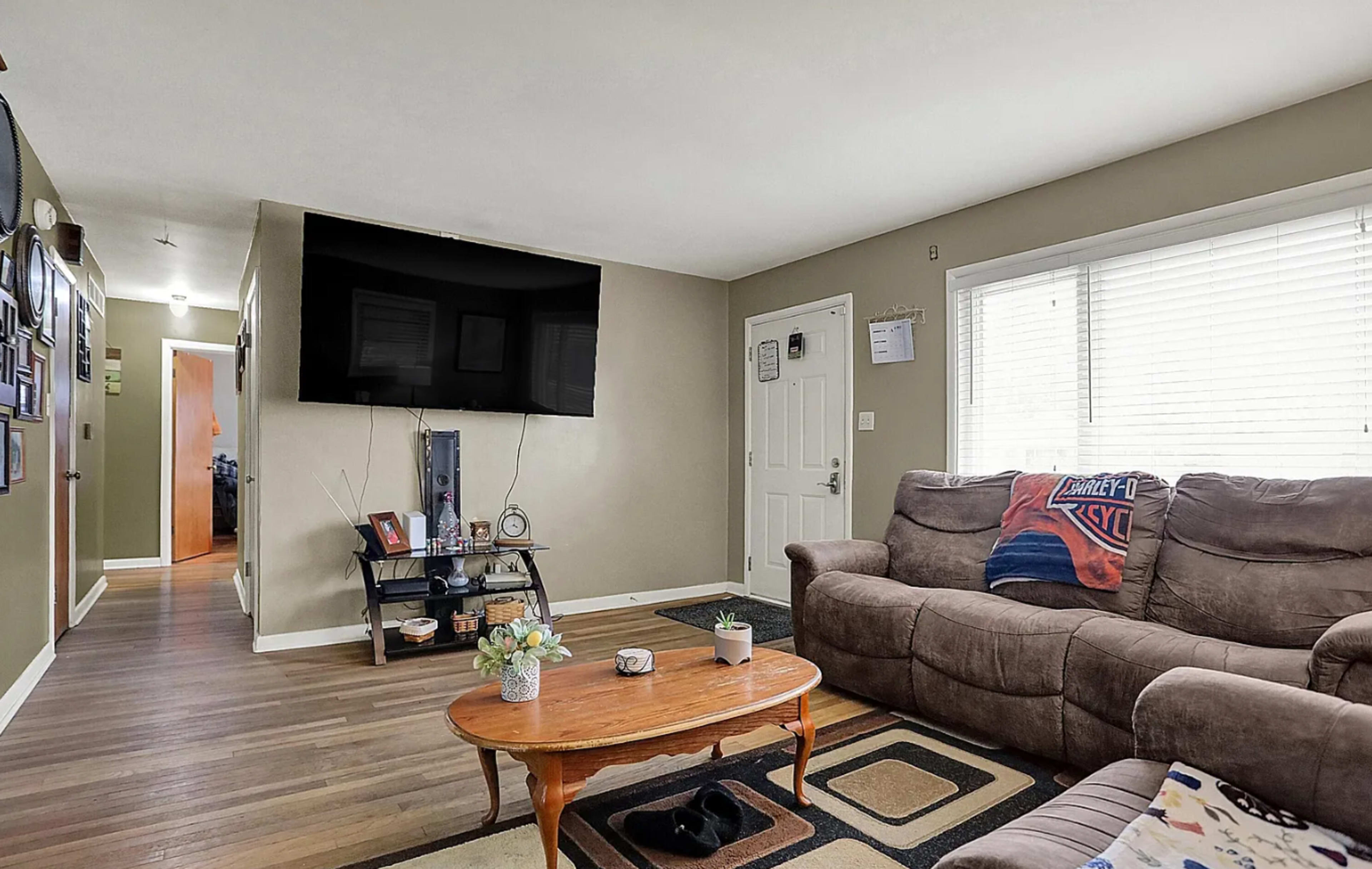 The image shows a living room with a large television mounted on the wall, a coffee table in front of a couch, and an entrance door visible in the background.