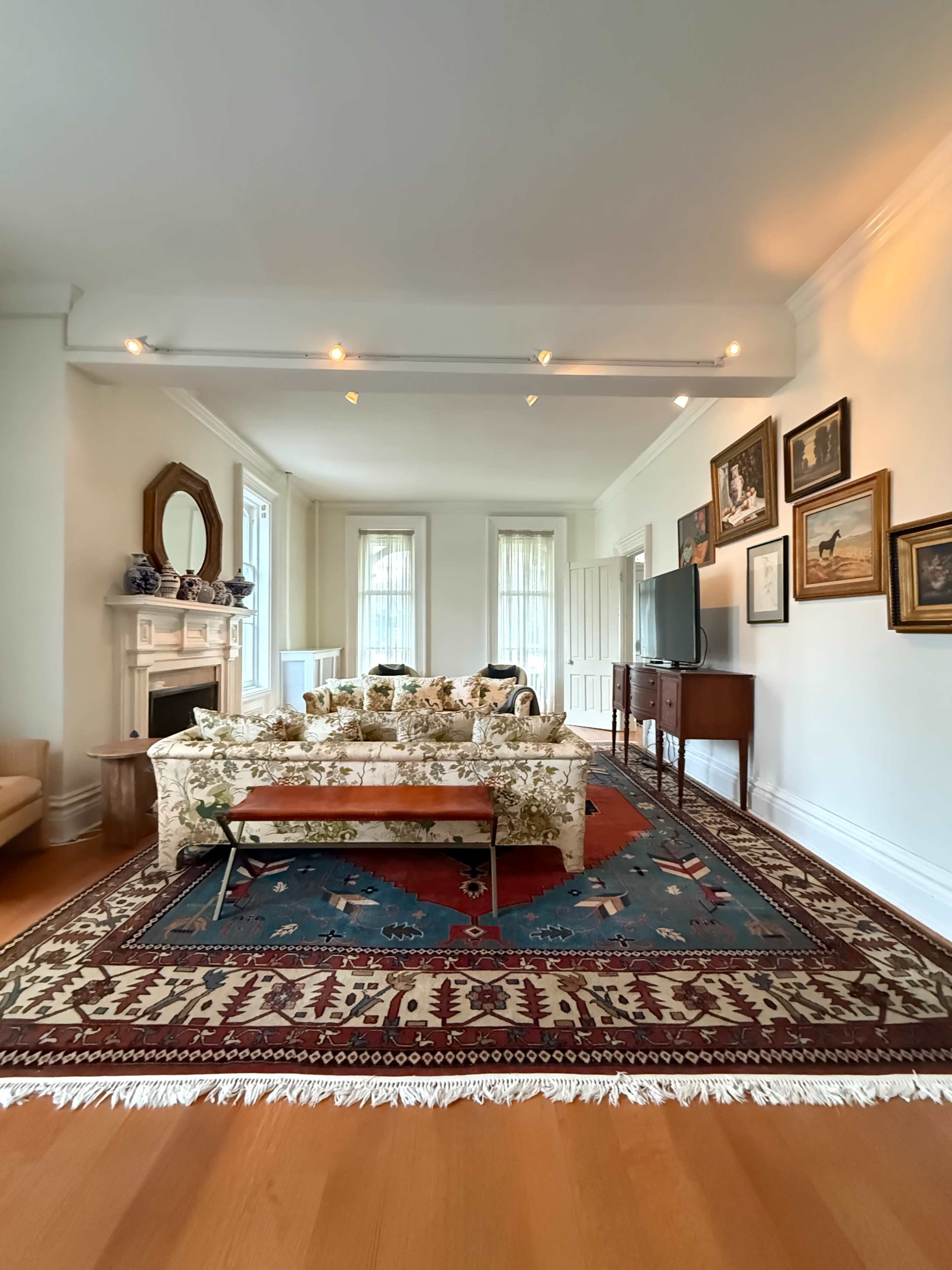 The image shows a spacious living room featuring a patterned rug, floral sofas, a wooden TV stand, and framed artwork on the walls.