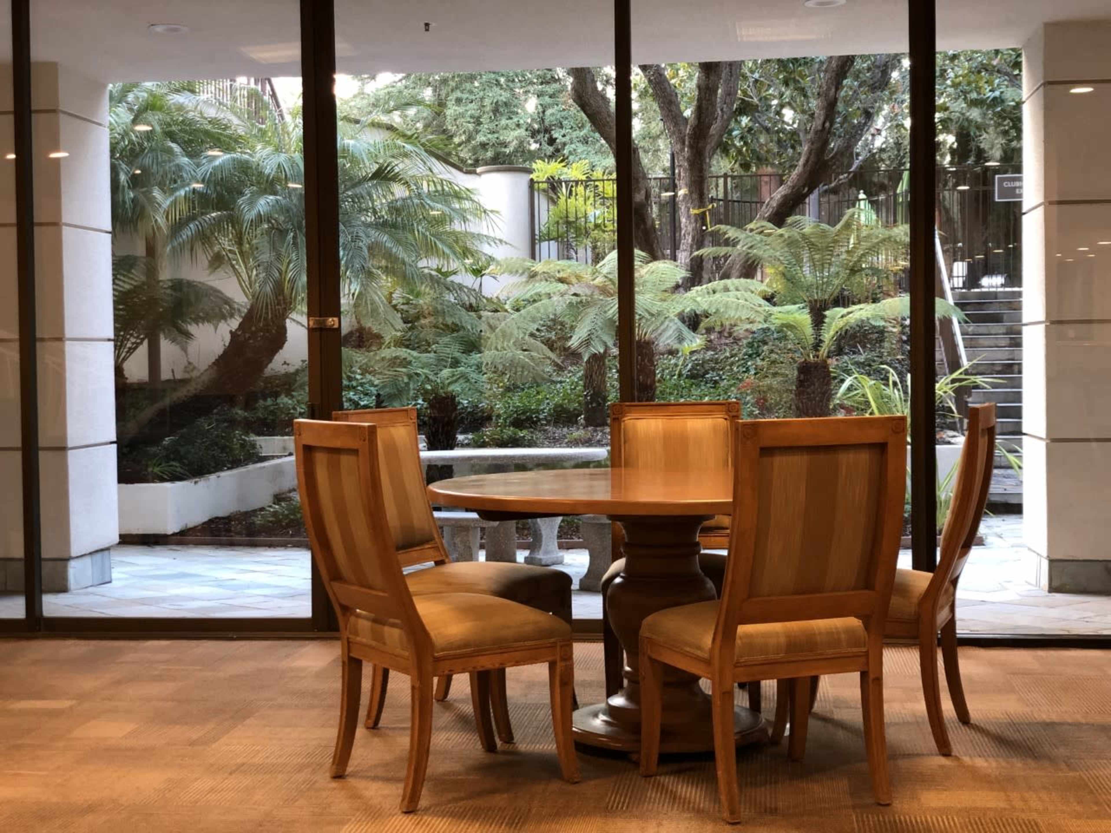 A wooden dining table with four chairs stands in front of large glass windows overlooking a garden with ferns and stone pathways.