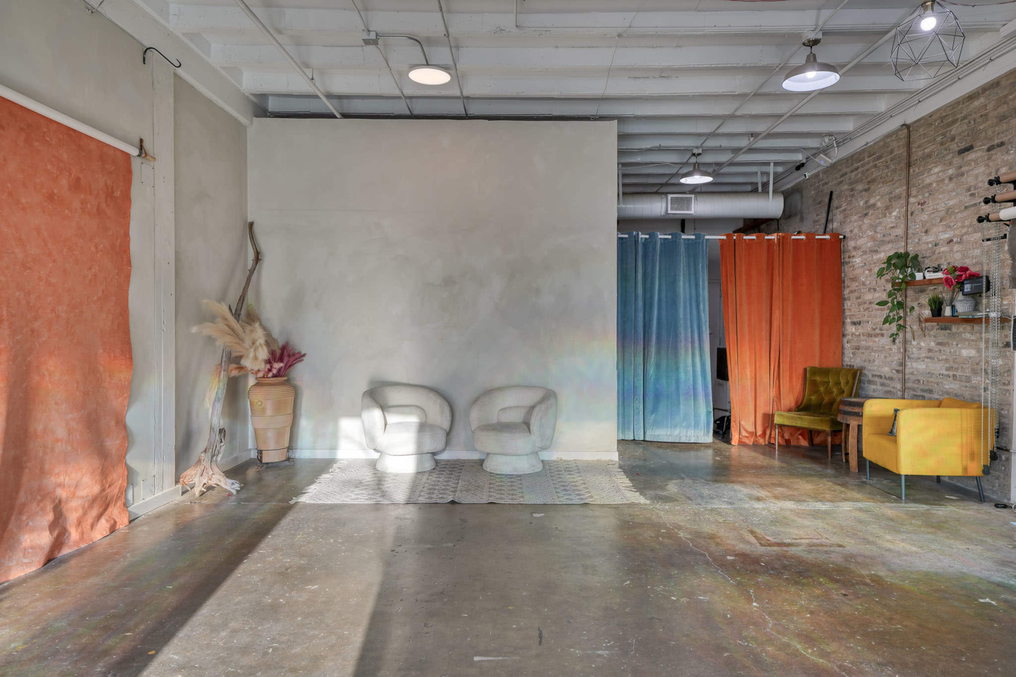 The image shows a spacious room featuring two armchairs, colorful fabric backdrops, and exposed brick walls.