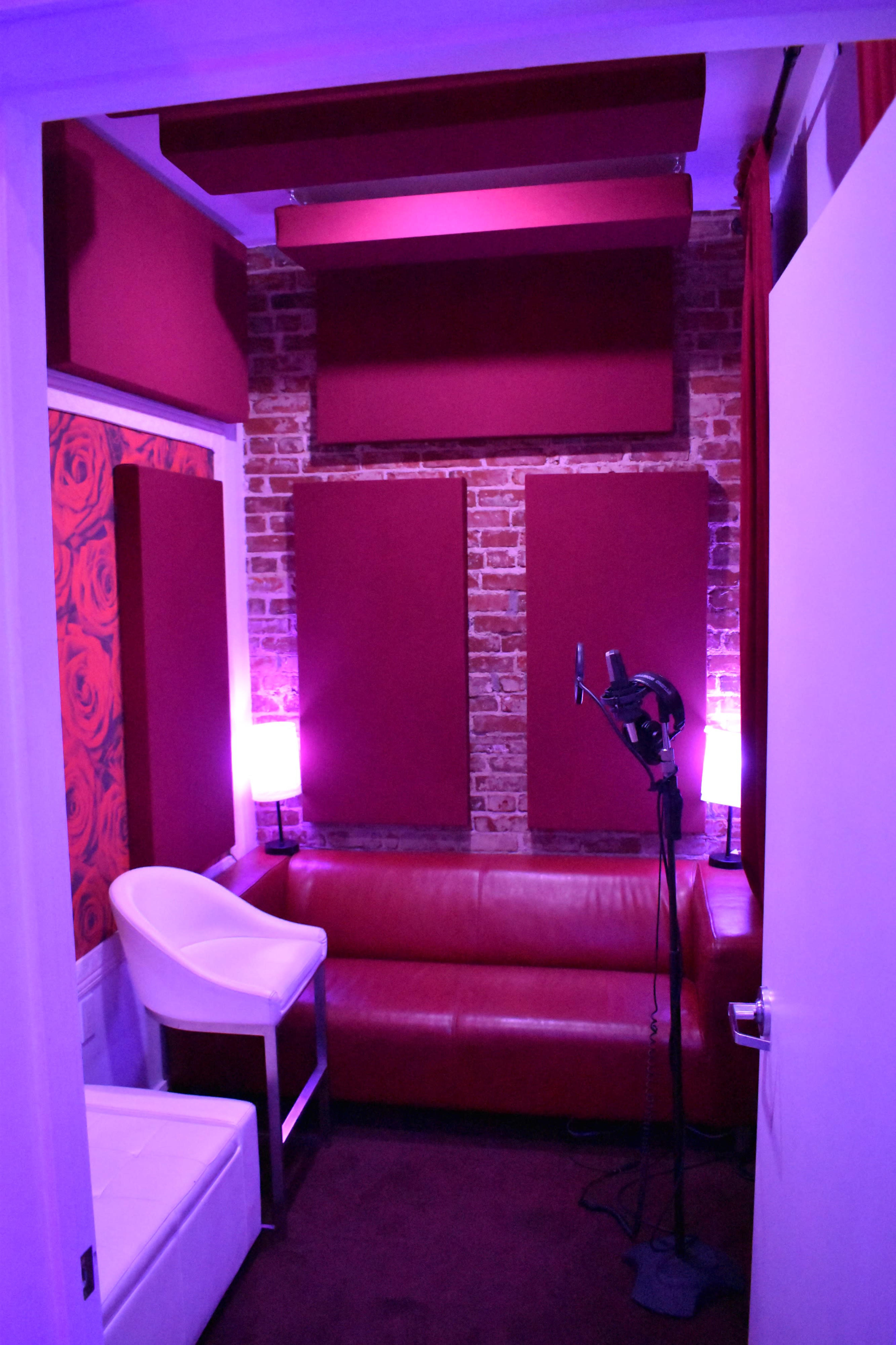 A small soundproof room with purple lighting, featuring a microphone stand, a leather sofa, and padded walls.