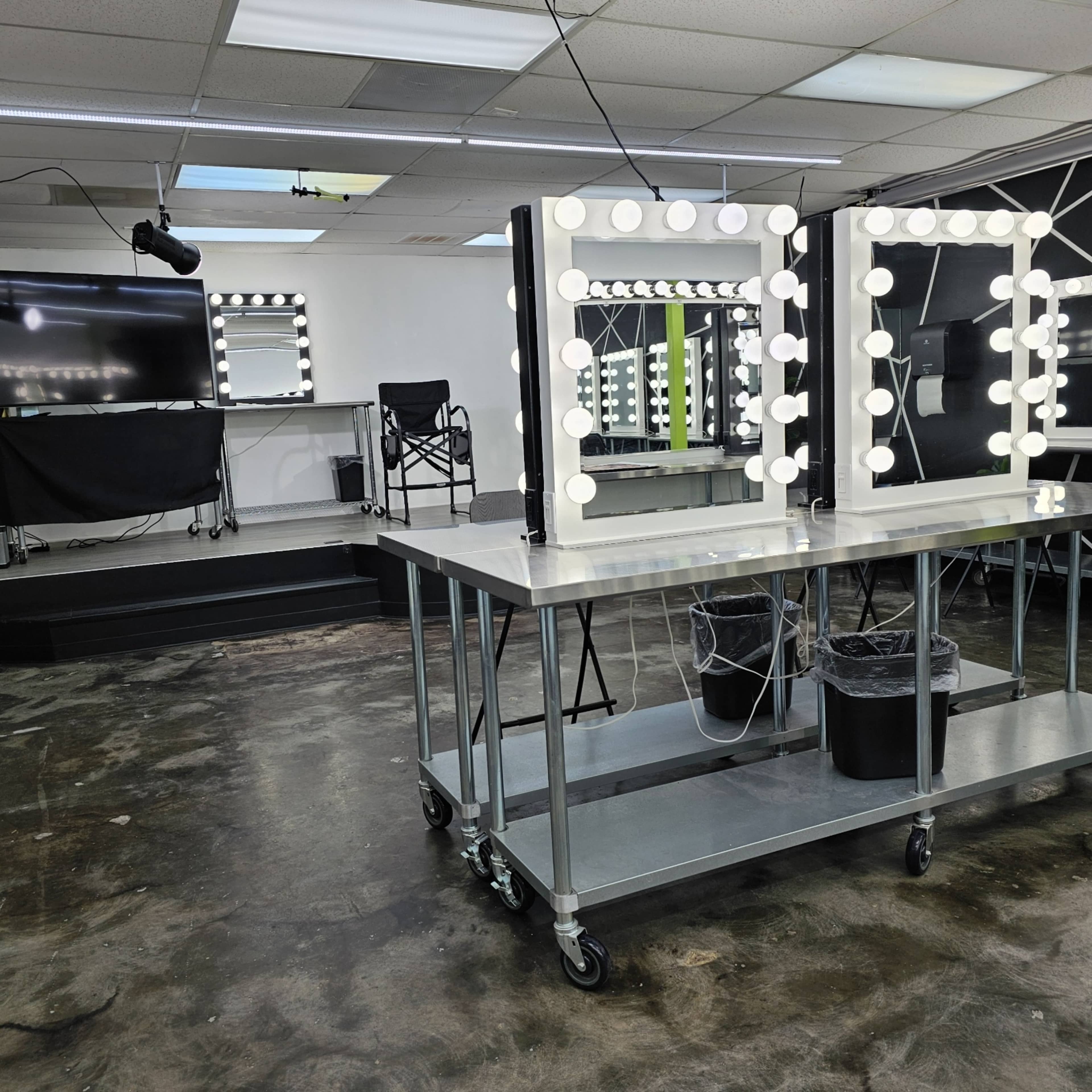 The image shows a well-lit makeup studio with two illuminated mirrors on tables, and a black chair and screen in the background.