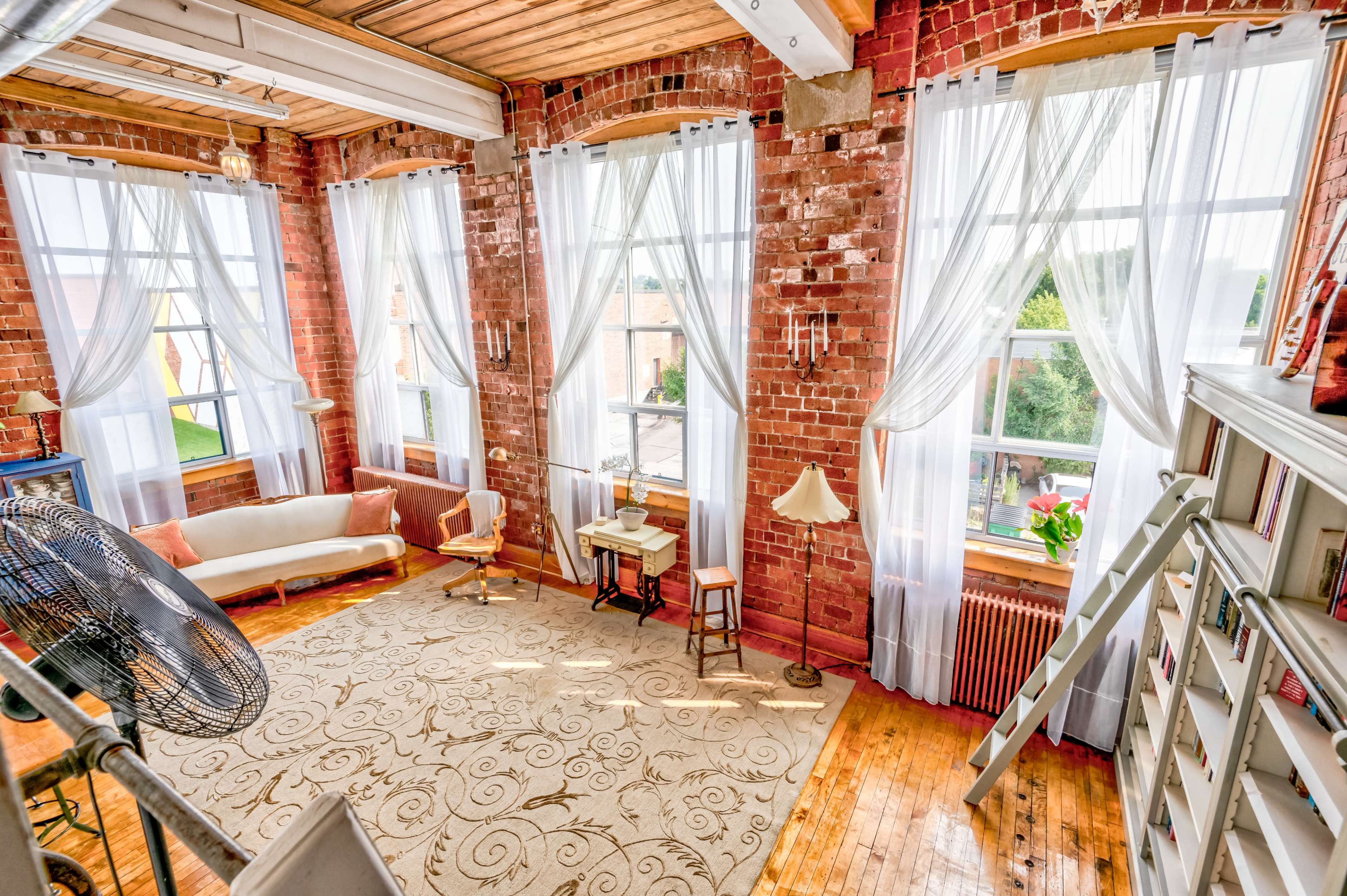 A spacious room with large windows dressed in sheer curtains, exposed brick walls, a light-colored sofa, a small desk with a lamp, and a decorative rug on a wooden floor.