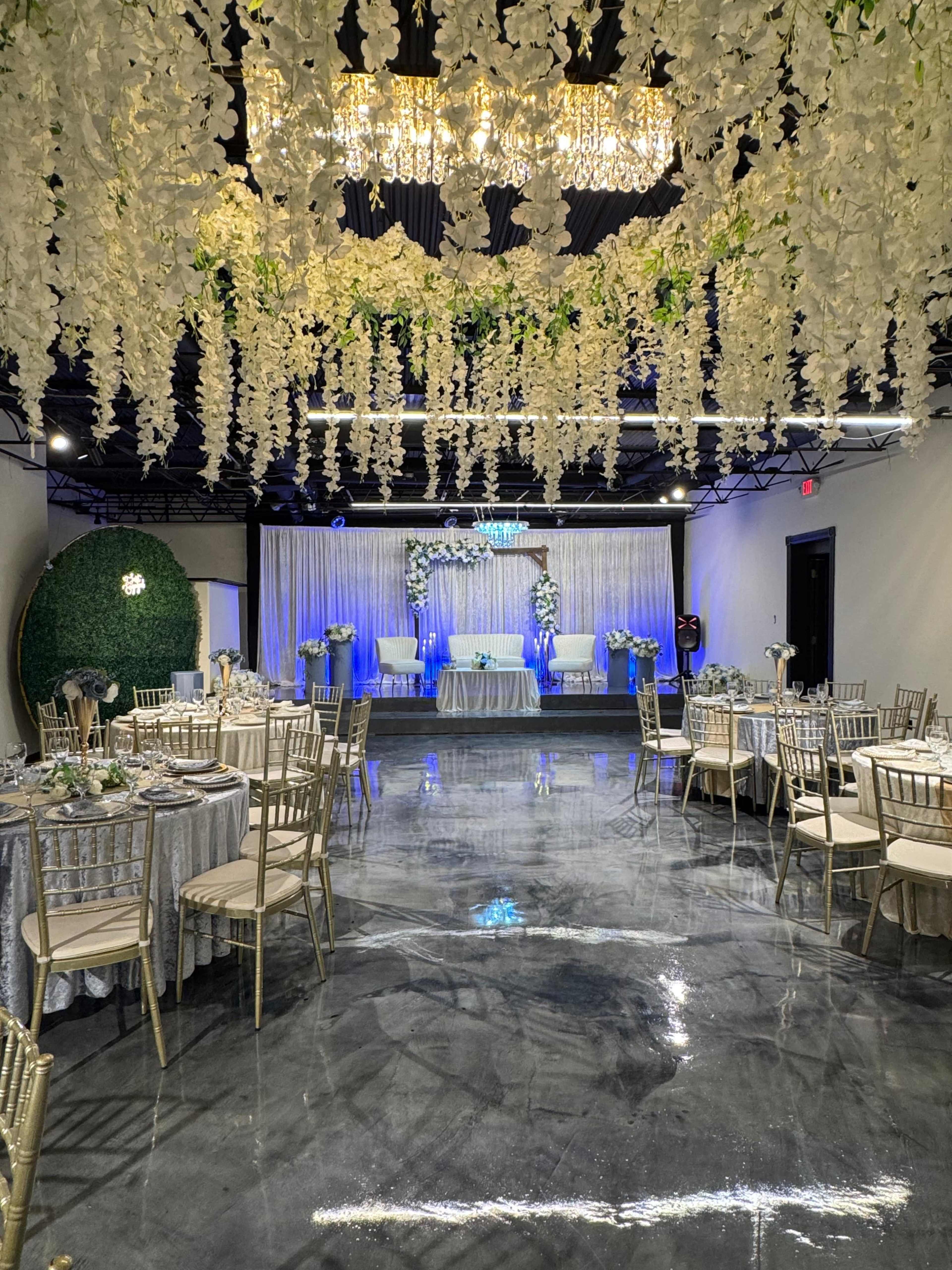 The image shows a decorated event space with elegant tables set for a celebration, featuring floral arrangements hanging from the ceiling and a backdrop with soft lighting.