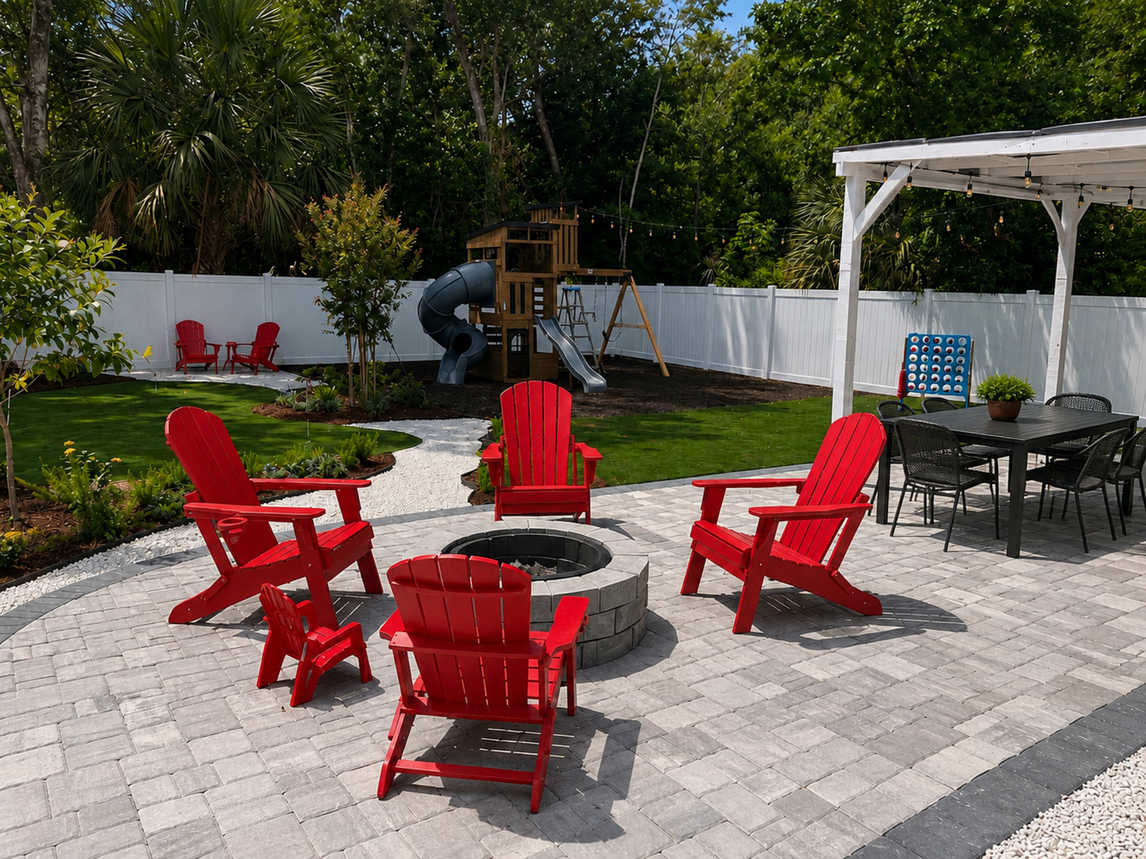 The image shows a backyard patio featuring red Adirondack chairs arranged around a circular stone fire pit, with a playground slide and play structure in the background.