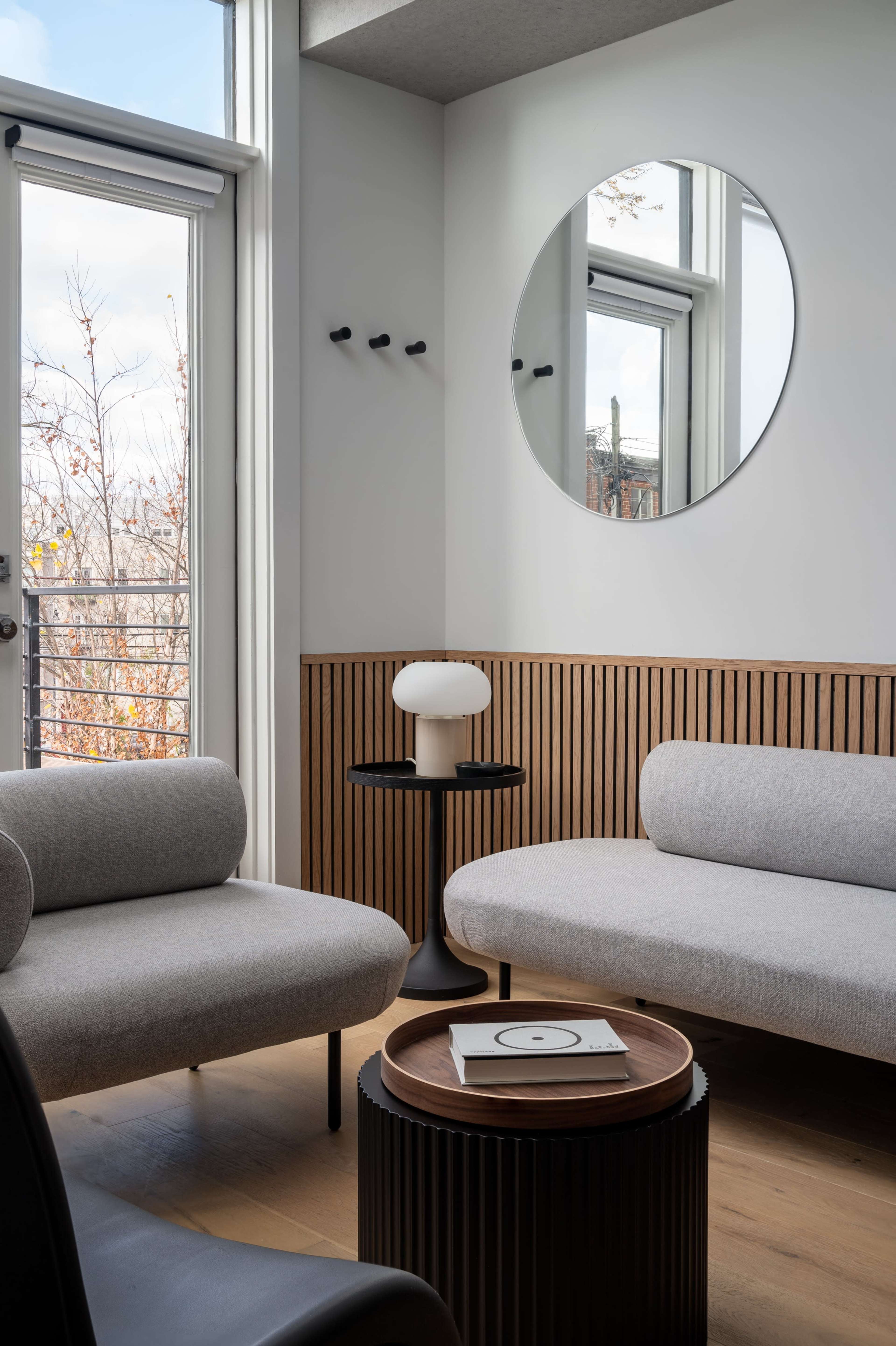 A modern living room features two gray sofas, a round mirror on the wall, and a small side table with a lamp.