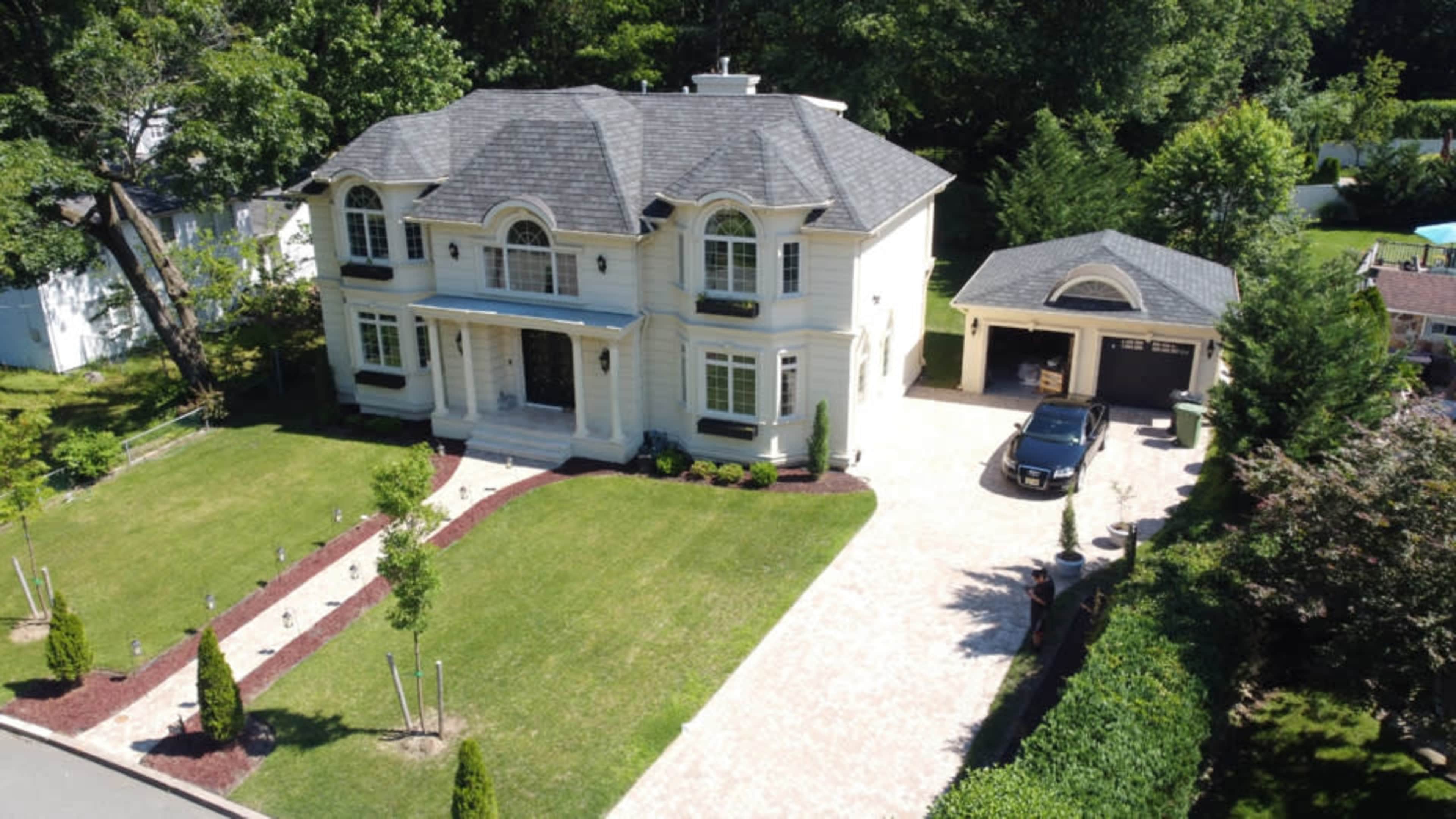 A large, two-story house with a gray roof and white exterior is surrounded by a manicured lawn and features a separate garage.