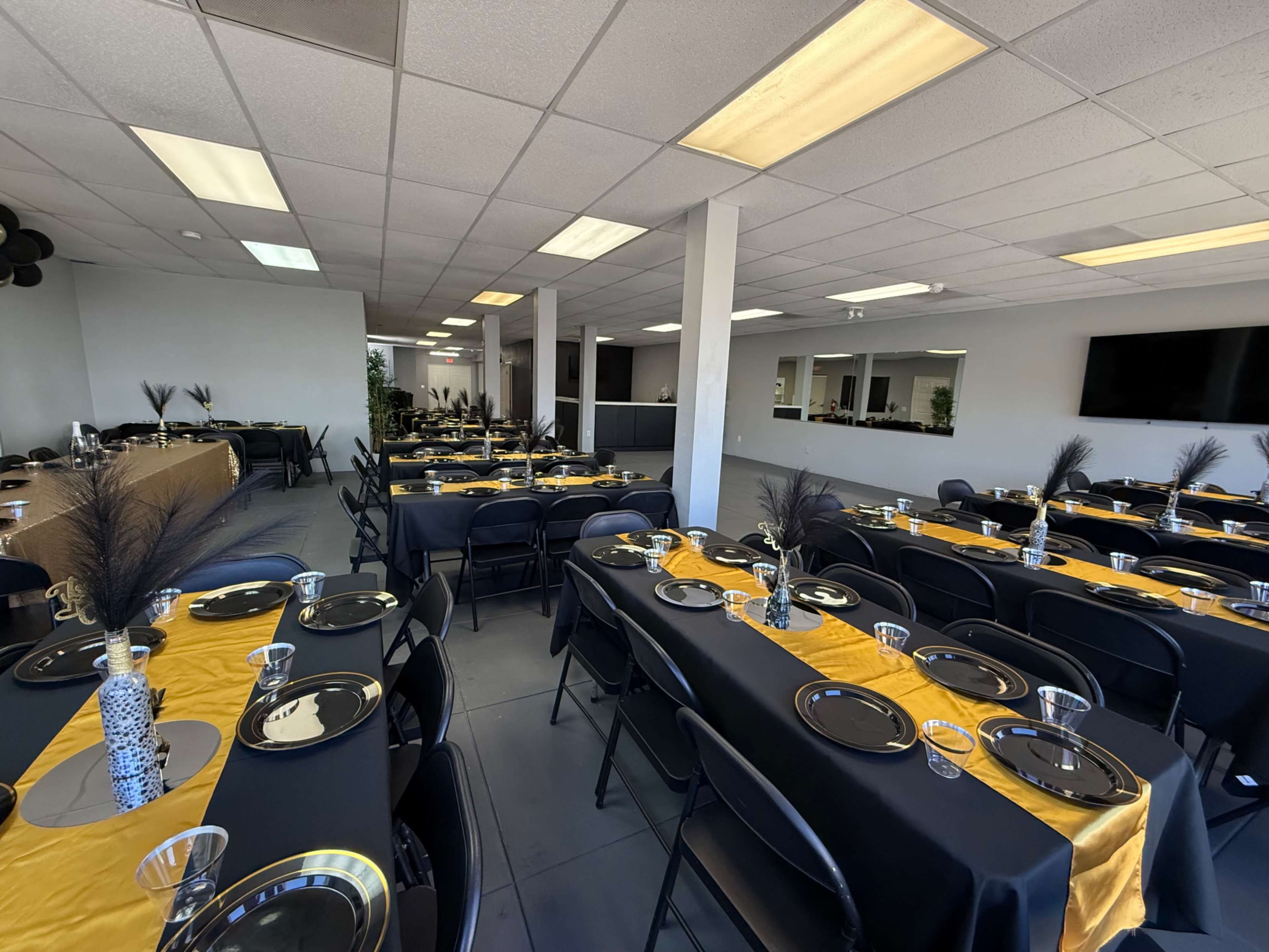 The image shows an event space set up with multiple tables covered in black and gold tablecloths, arranged for a gathering.