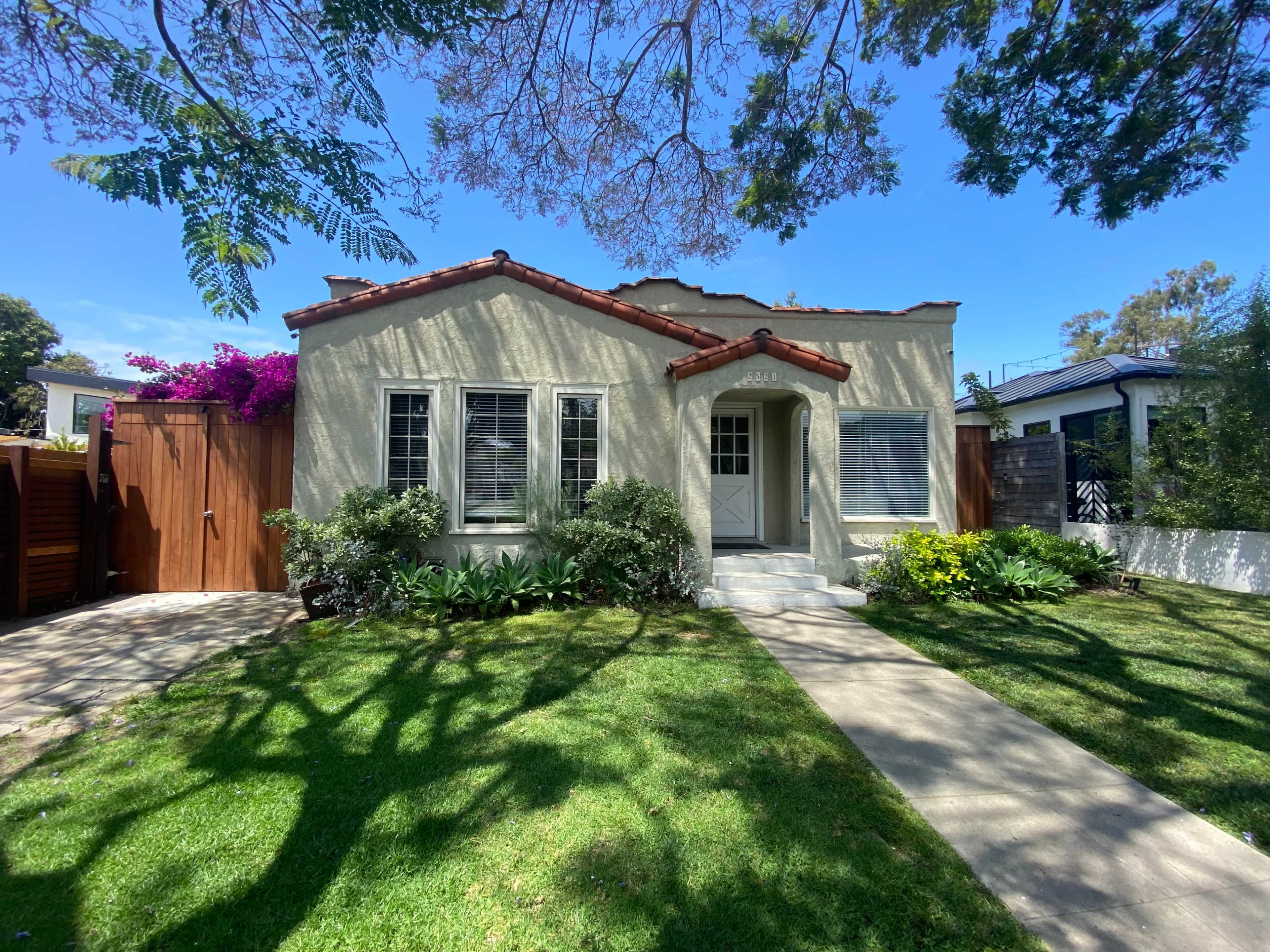 The image shows a single-story house with a light-colored exterior, a red-tiled roof, and a well-maintained front yard featuring shrubs and flowering plants.