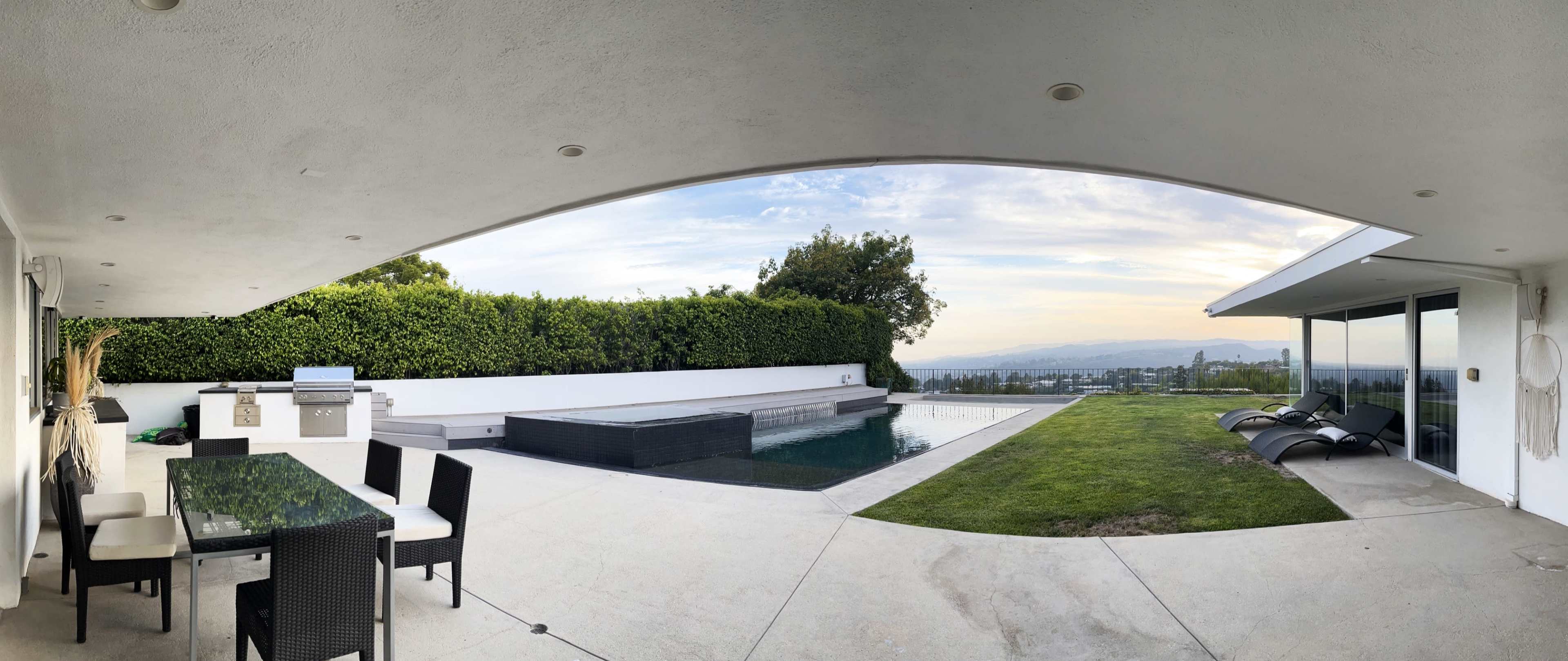 The image shows a modern outdoor patio area with a dining set, a barbecue grill, a pool, and a view of a landscaped yard and distant hills under a partly cloudy sky.