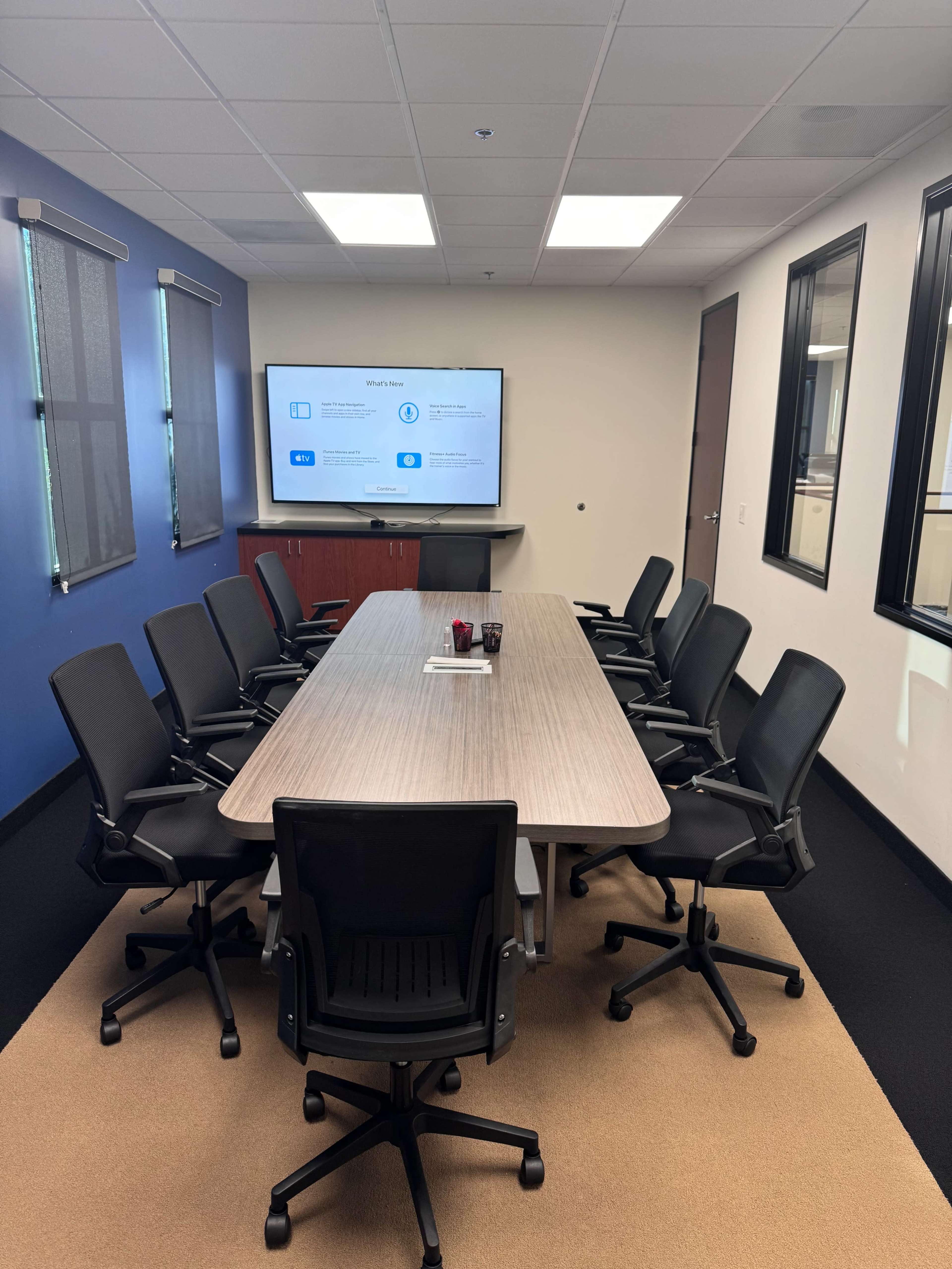 A modern conference room features a long table surrounded by black chairs, with a large screen displaying a presentation on the wall.