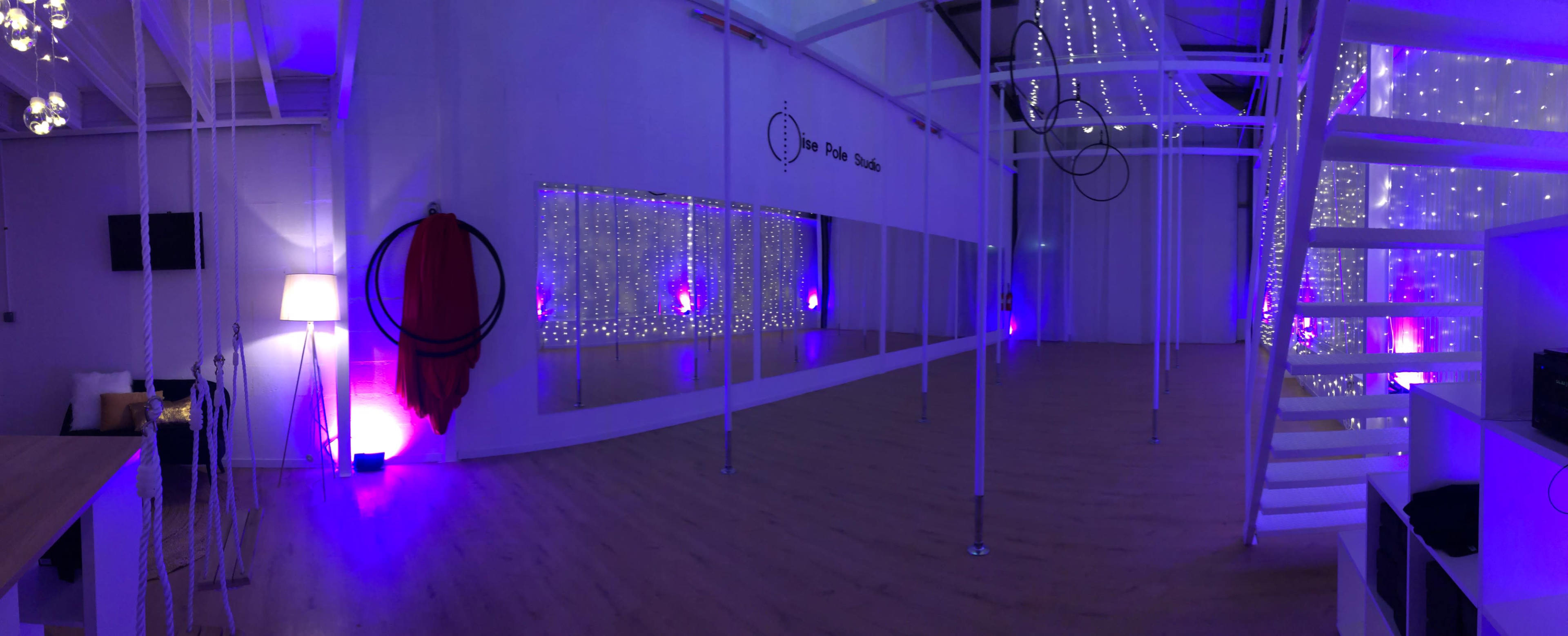 A spacious studio featuring mirrored walls, a wooden floor, and soft purple lighting with aerial silks and hoops hanging from the ceiling.
