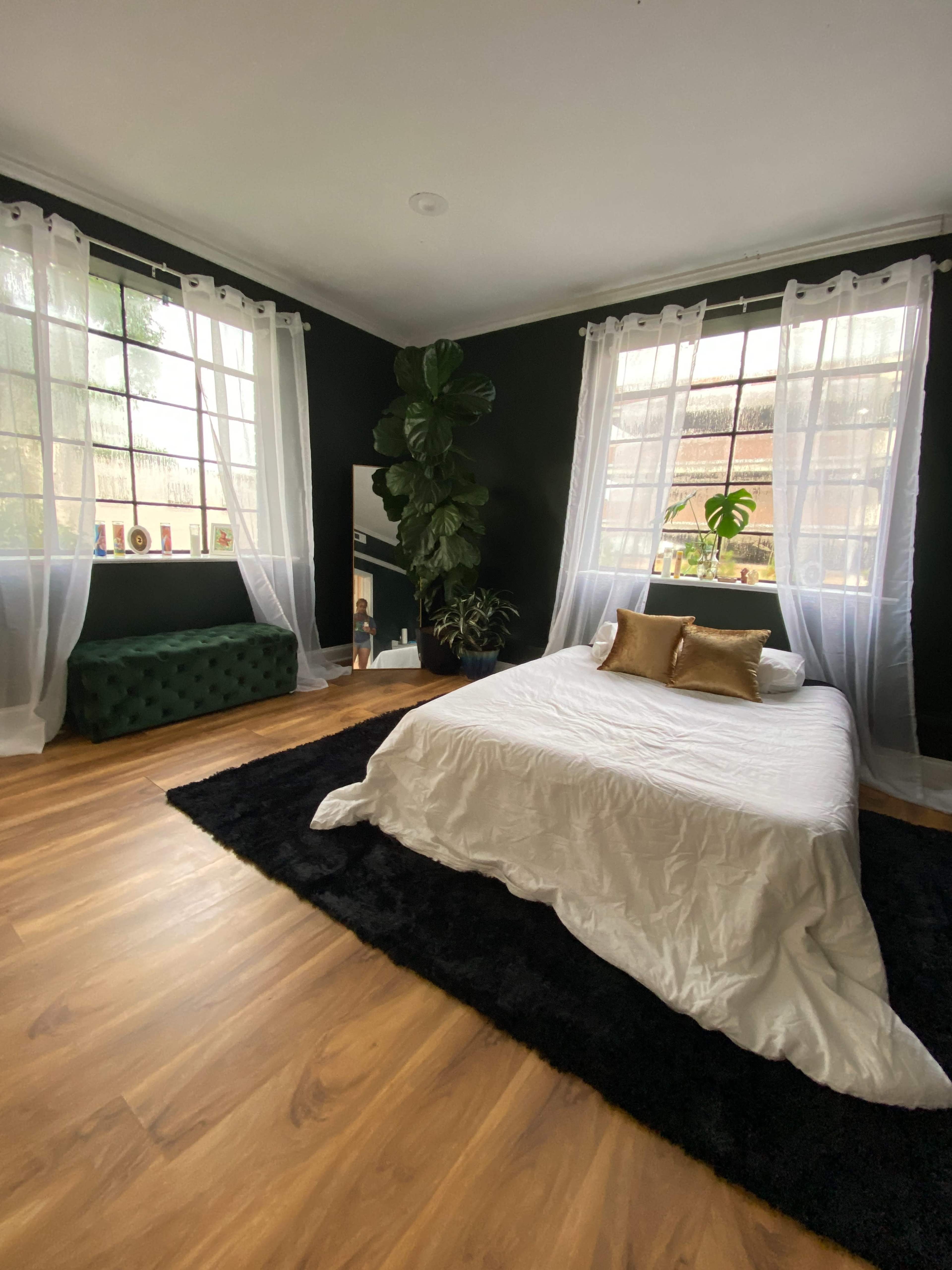 The bedroom features a bed with white bedding and gold cushions, surrounded by dark green walls, large windows, and a faux fur rug.