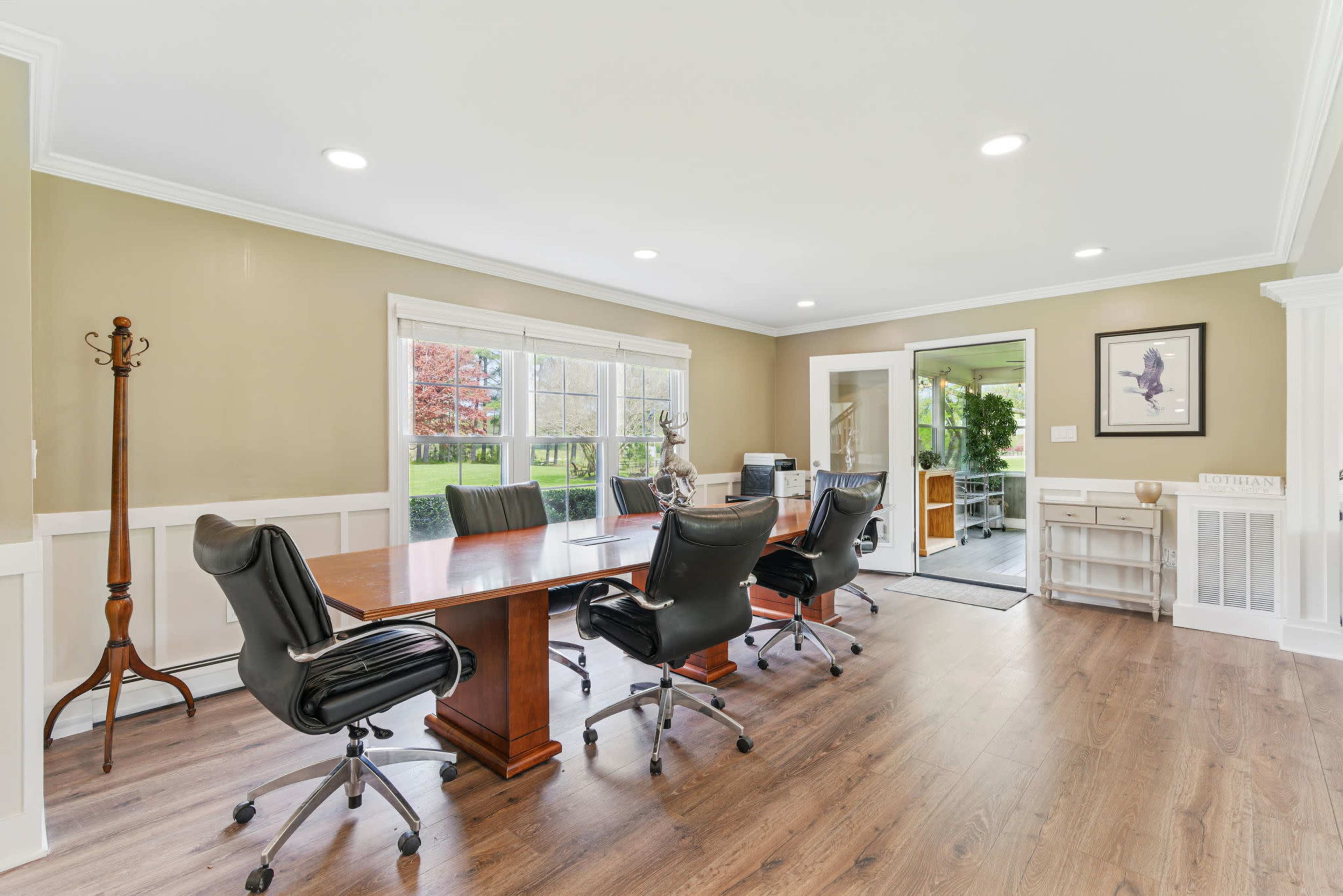The image depicts a conference room featuring a large wooden table surrounded by several black leather chairs, with windows providing a view of greenery outside.