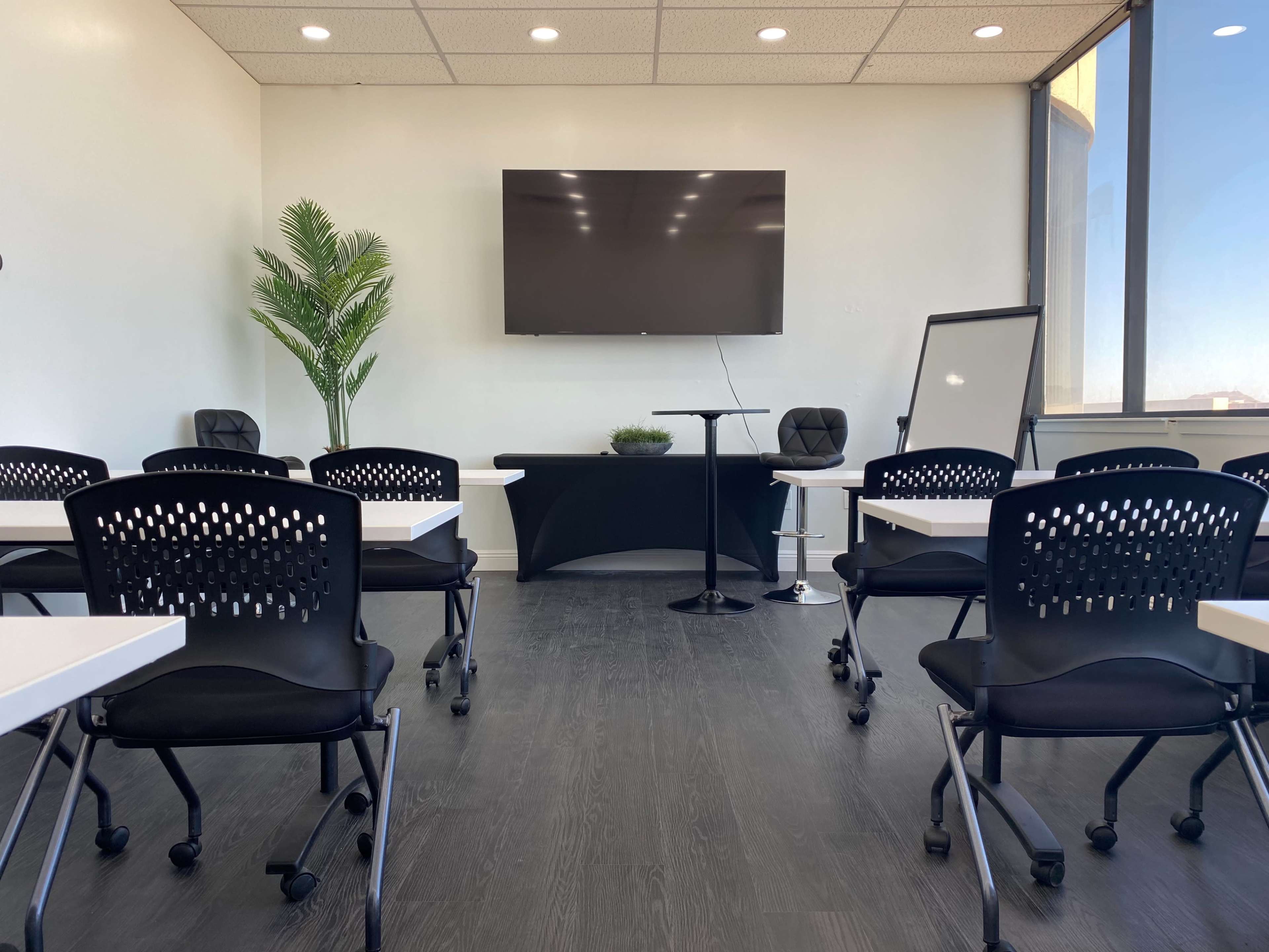 A modern classroom is set up with several black chairs and tables, facing a large wall-mounted television and a potted plant in the corner.