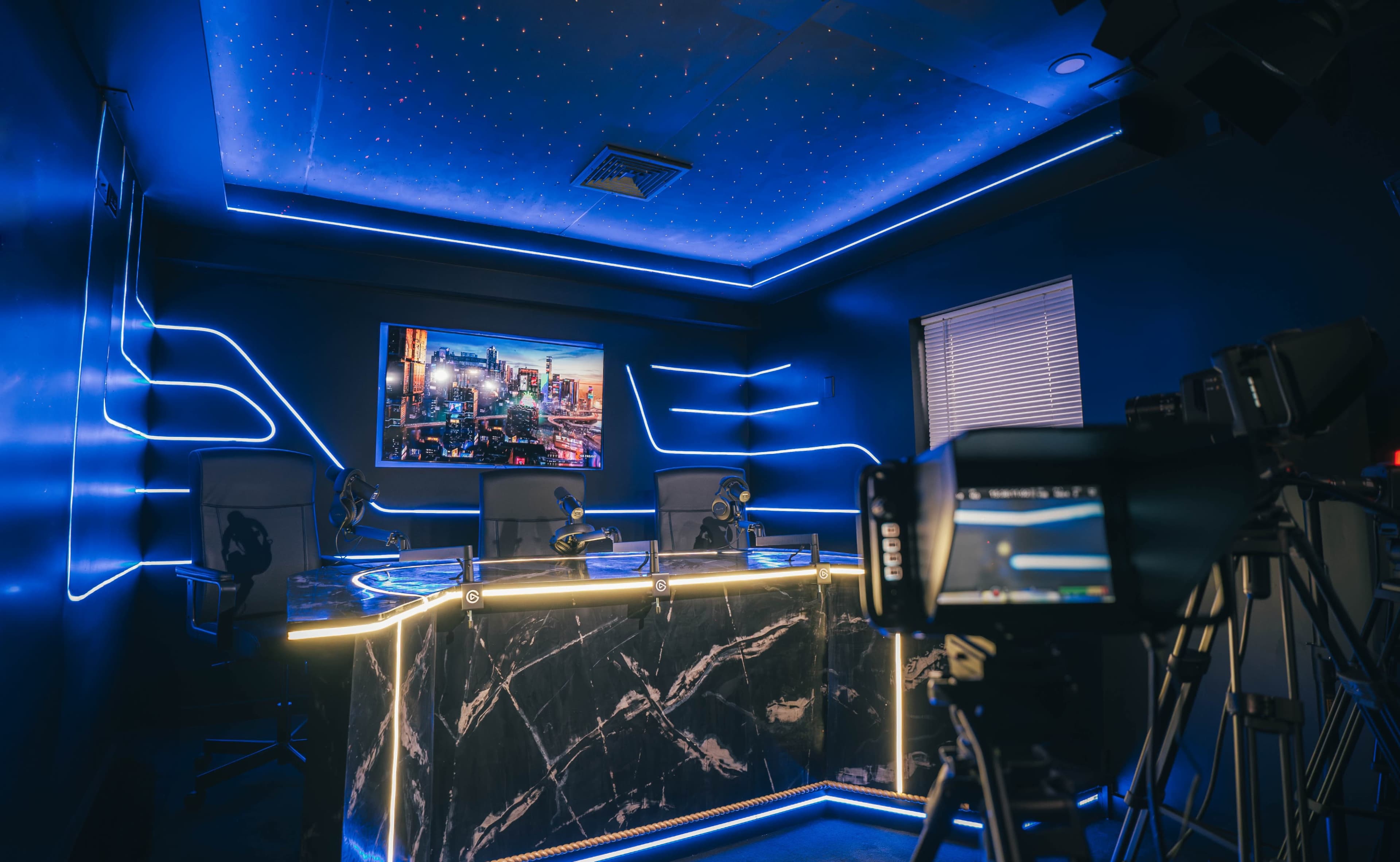 A modern studio setup featuring a sleek desk with microphones, a large screen displaying a cityscape, and ambient blue lighting.