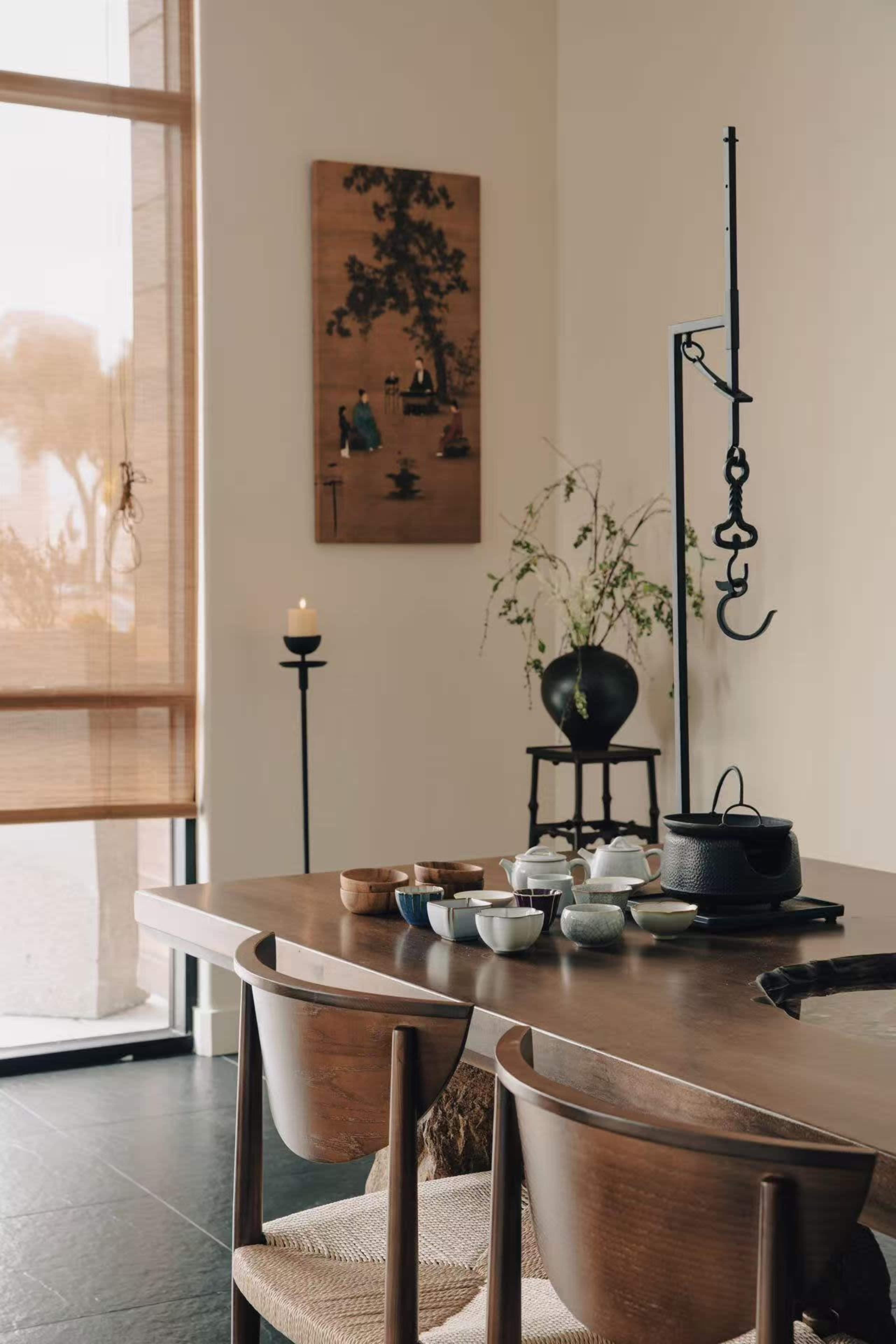 A wooden dining table set with various bowls and a teapot is positioned in a room with natural light, featuring a wall art piece and a potted plant nearby.