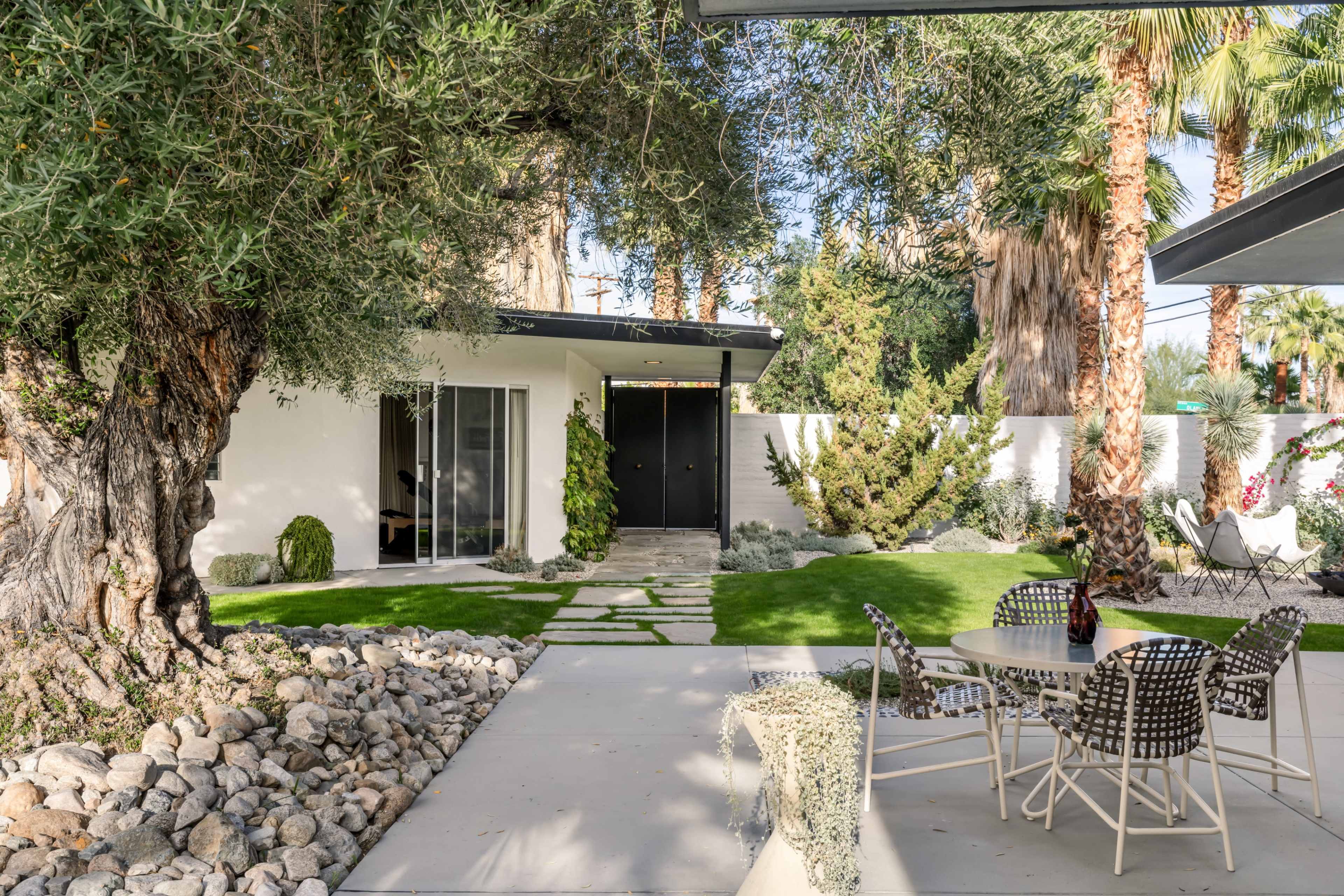 The image shows a modern outdoor space with a patio area featuring a table and chairs, surrounded by landscaped greenery and palm trees.