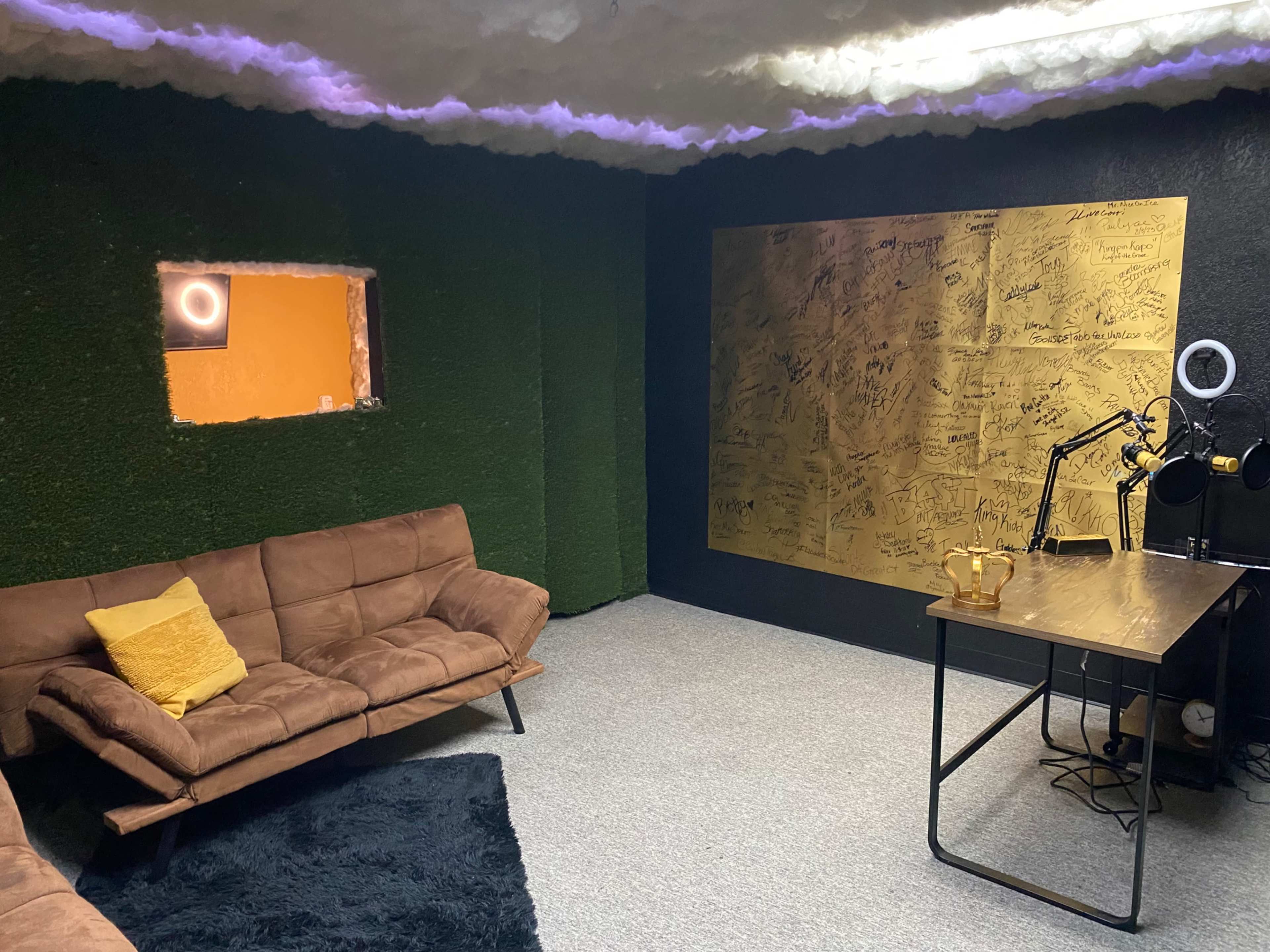 The image shows a modern room with a green moss-covered wall, brown couches, a gold wall covered in signatures, and a desk with a microphone setup.