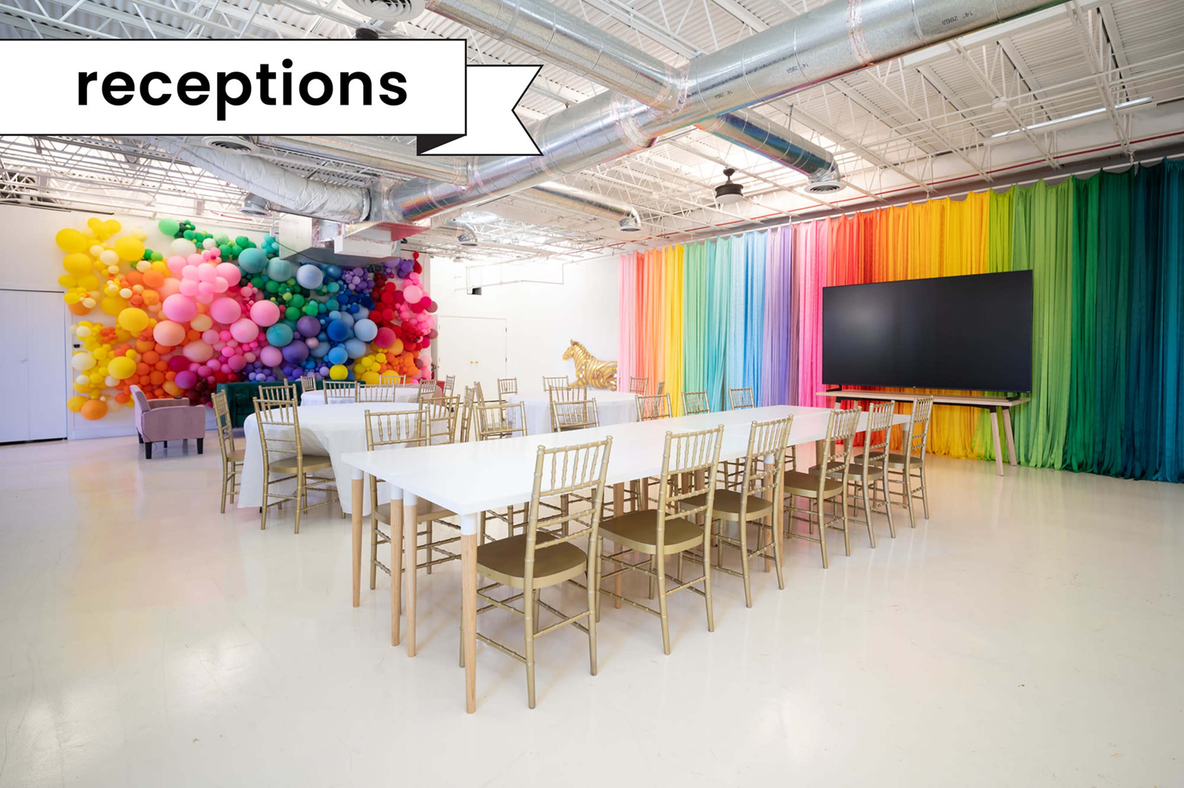 A brightly colored reception area features a long white table surrounded by gold chairs, with a vibrant balloon arch and a large screen at one end of the room.