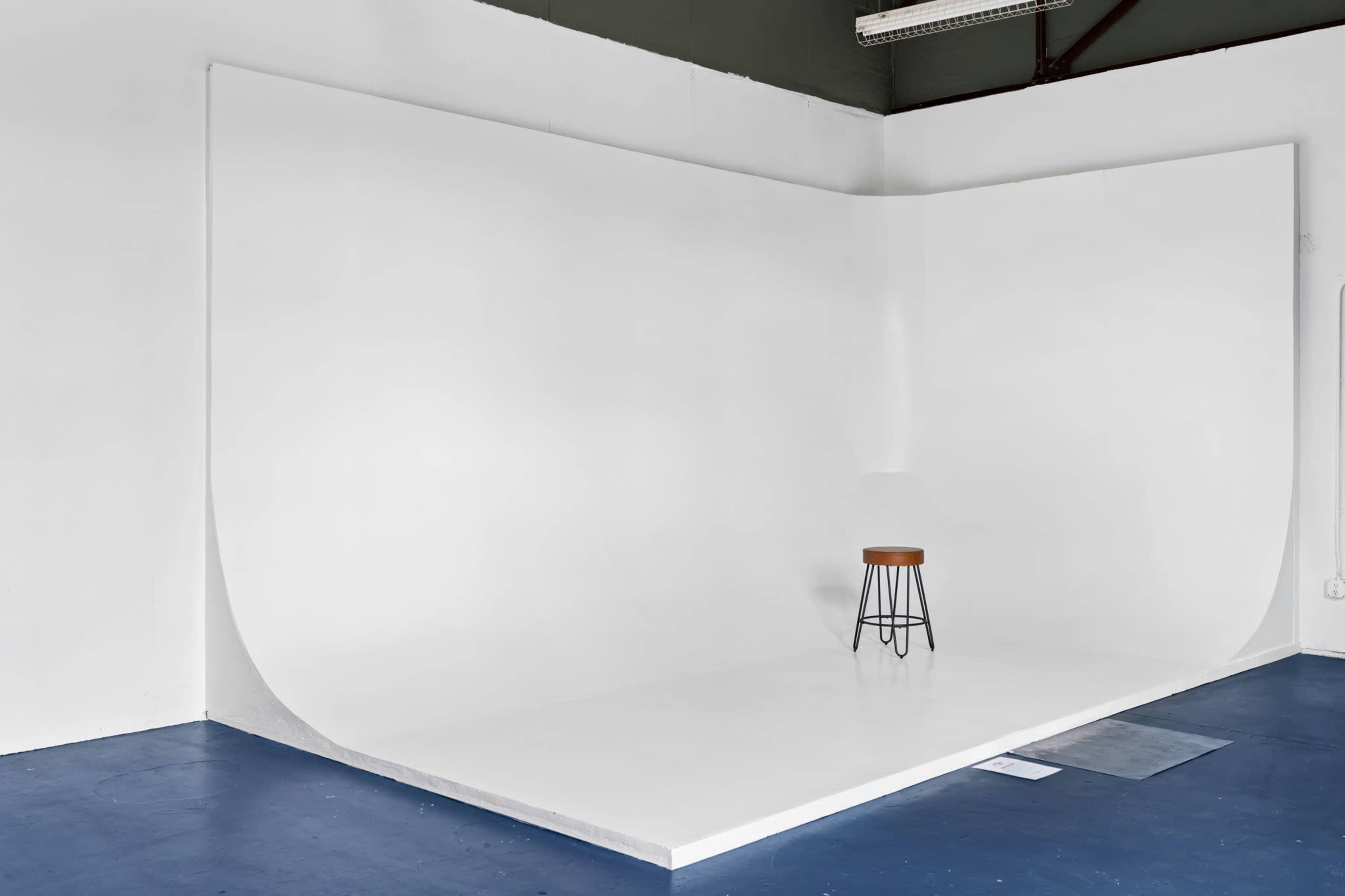 A blank studio space features a seamless white backdrop and a single wooden stool on a blue floor.