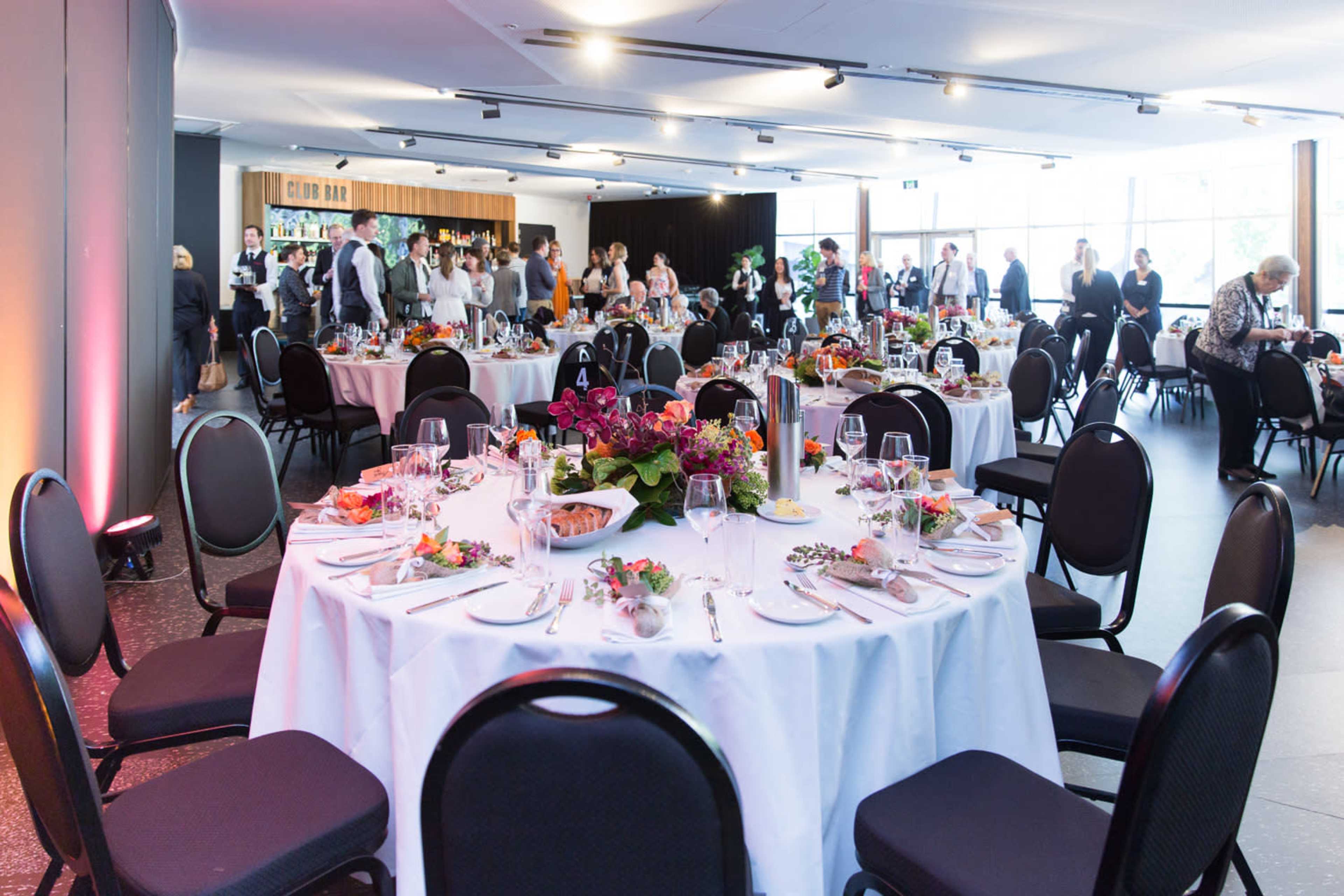 A banquet hall is set with elegantly arranged tables, featuring white tablecloths and floral centerpieces, while guests mingle in the background.