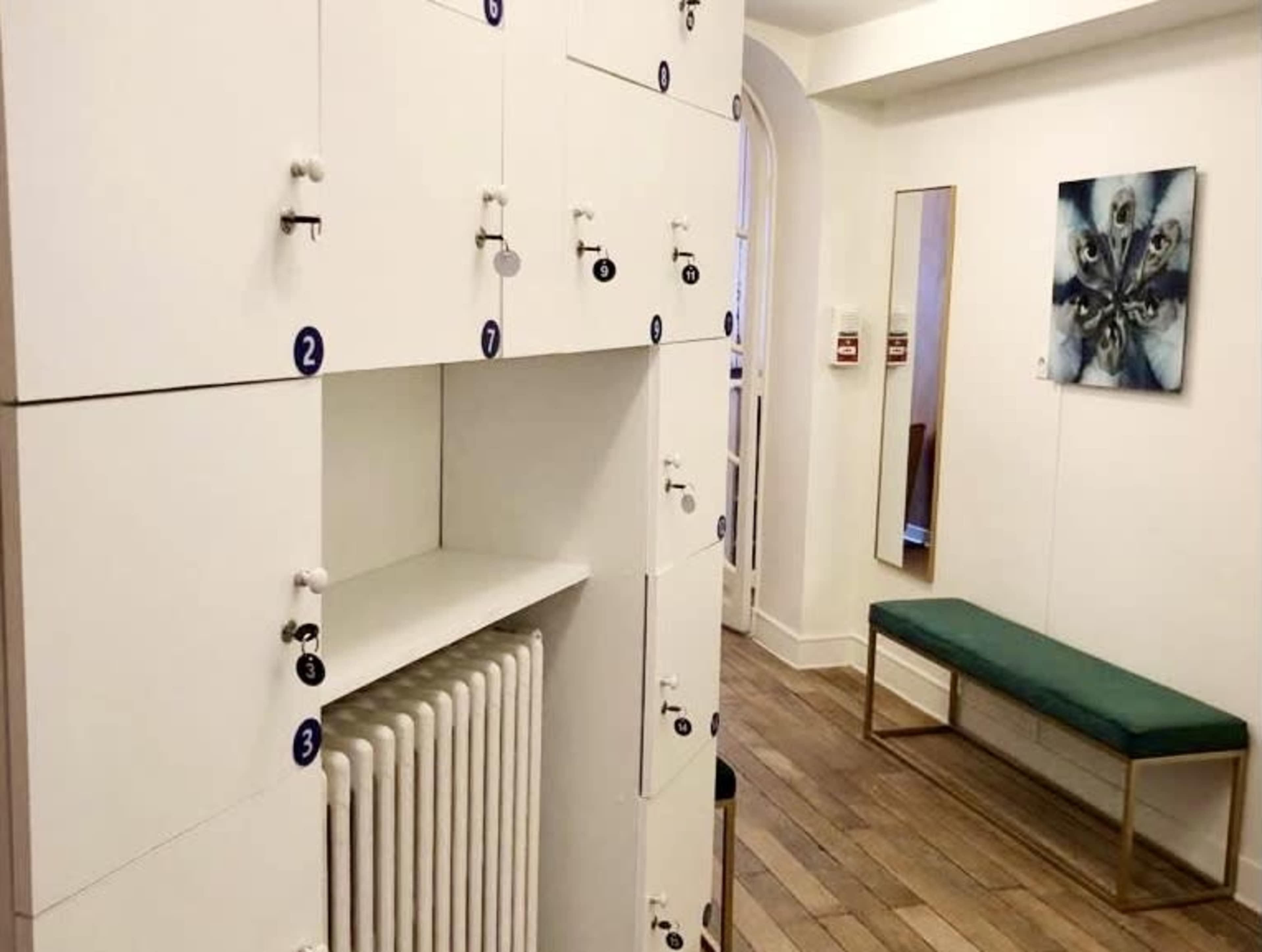 A corridor with white lockers numbered 1 to 3 on one side, a green bench, a mirror, and a floral painting on the opposite wall.