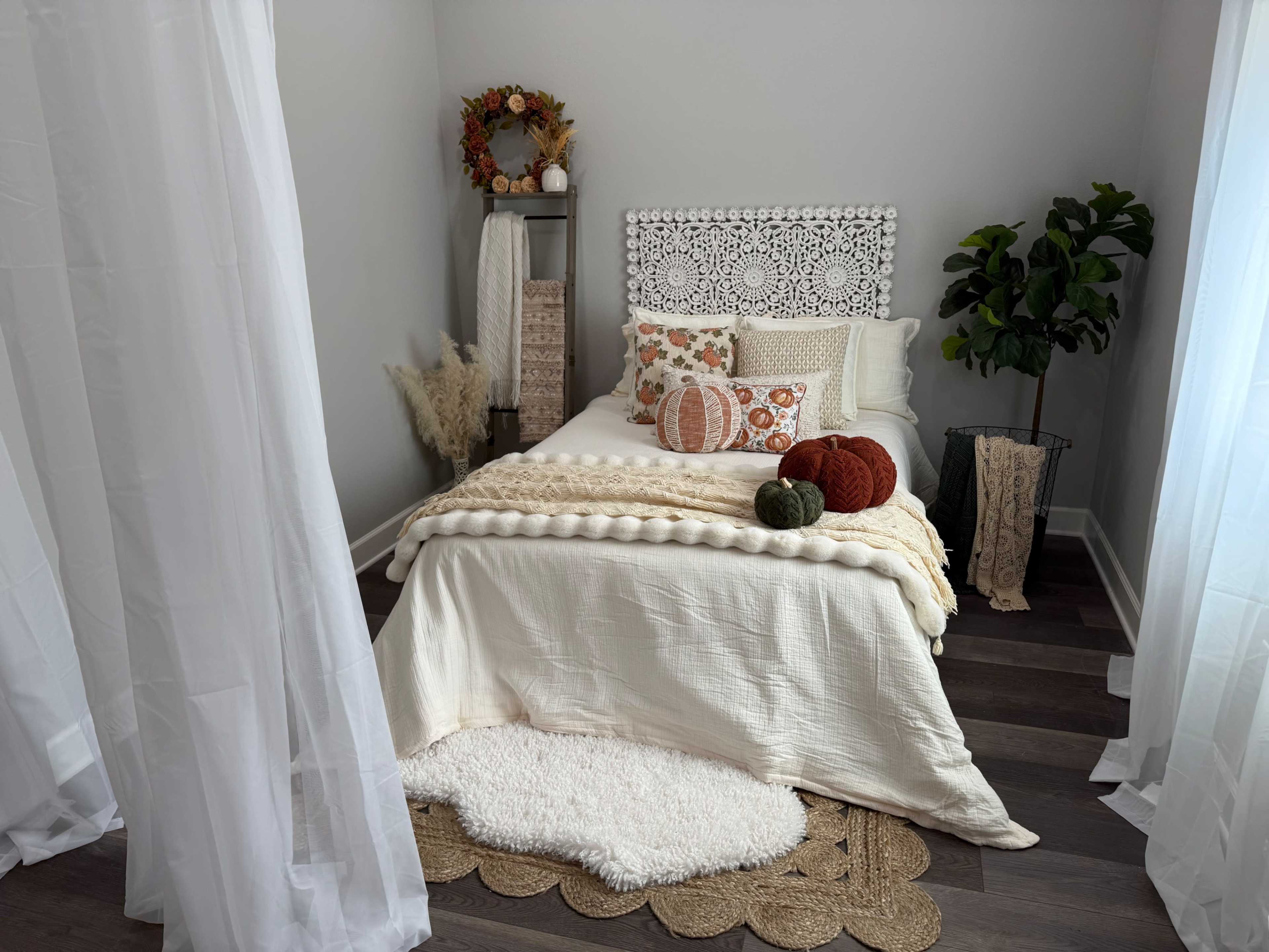 The image shows a cozy bedroom with a bed adorned with decorative pillows, a textured blanket, and pumpkins, surrounded by sheer curtains and a potted plant.