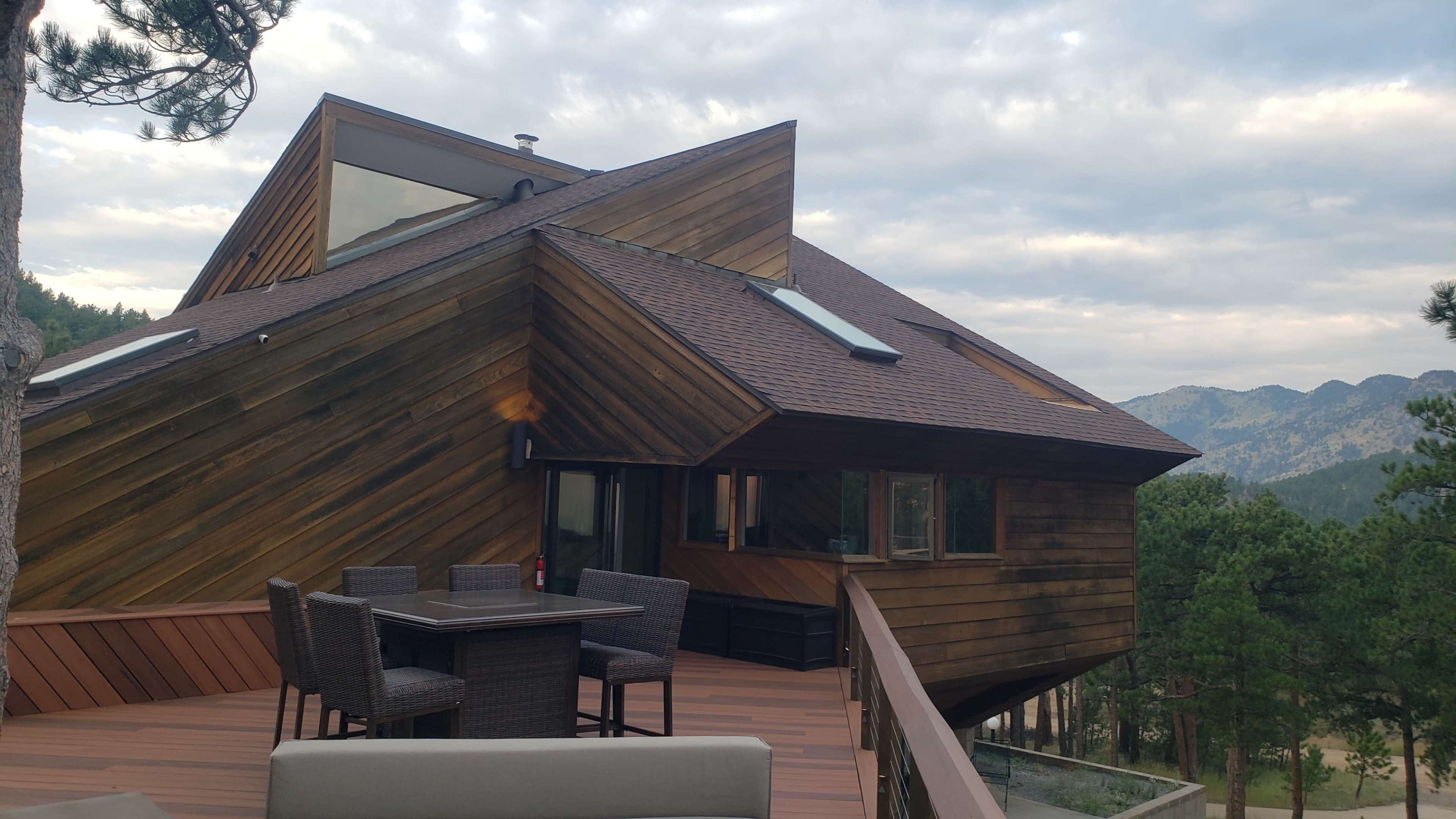 A modern wooden house with multiple roof angles overlooks a deck furnished with a dining set and surrounded by pine trees and mountainous terrain.