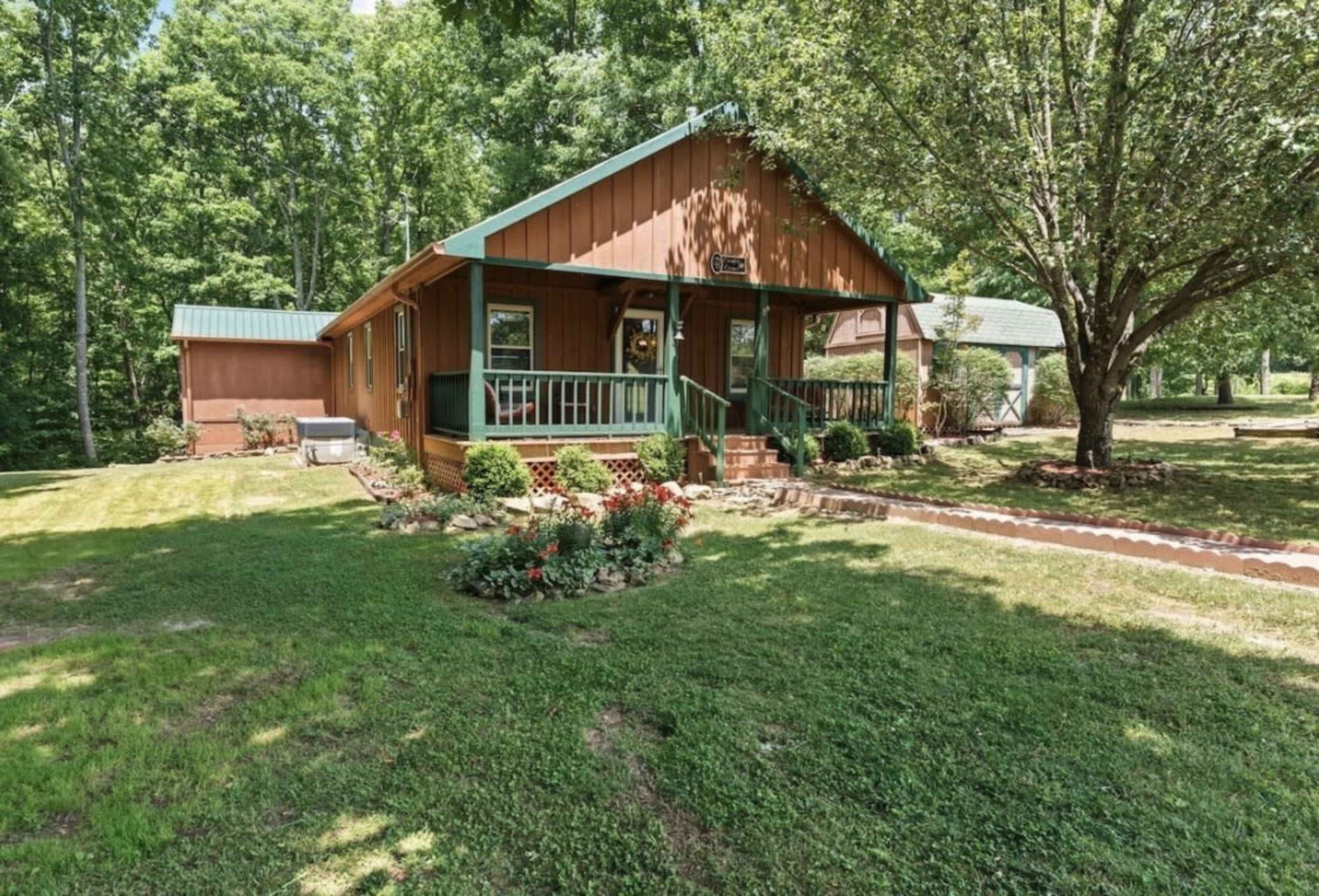 The image shows a brown wooden house with a green roof, surrounded by a green lawn and trees.