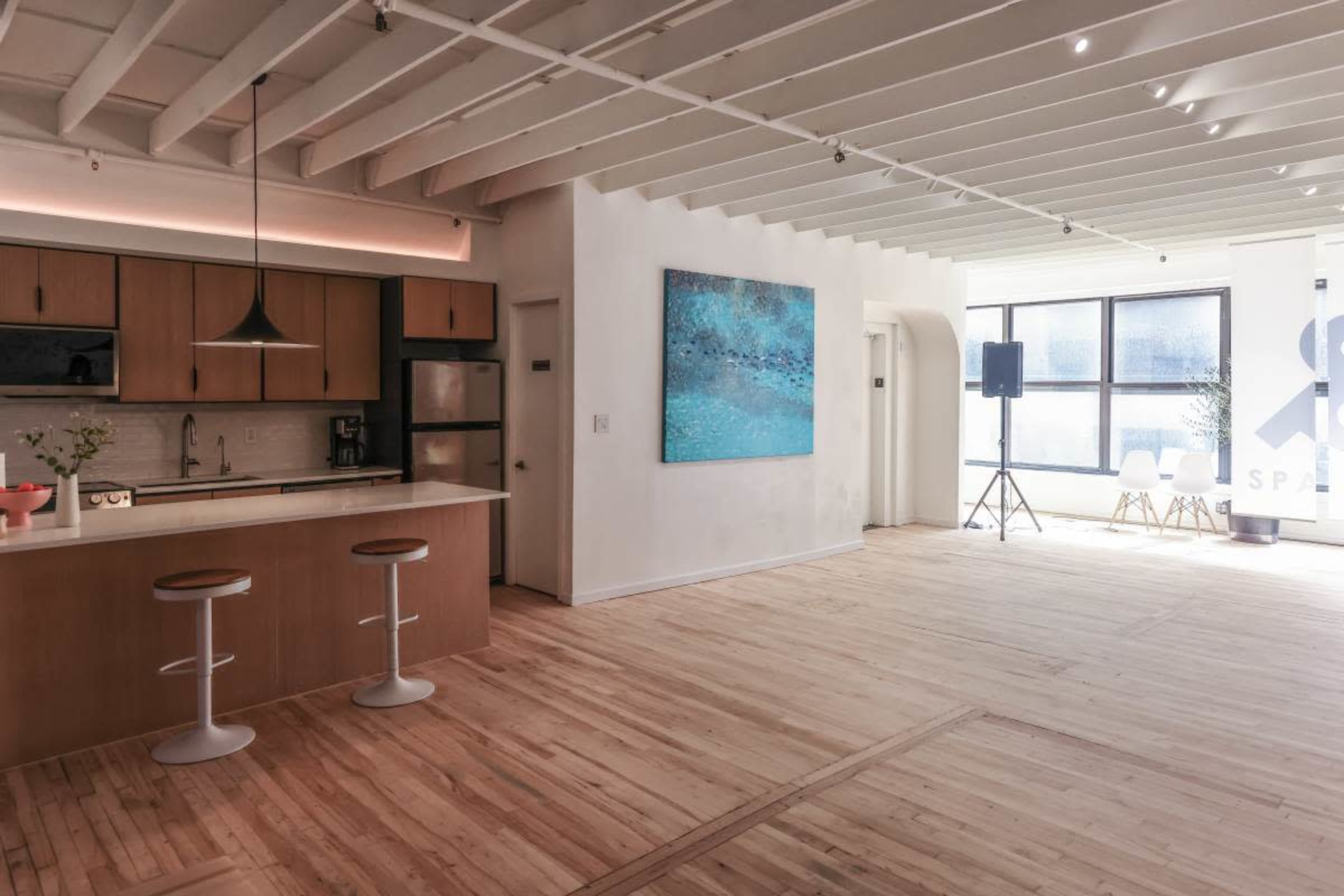 The image shows a spacious, modern kitchen and living area featuring a large wall painting, wooden floors, and two bar stools at the kitchen counter.
