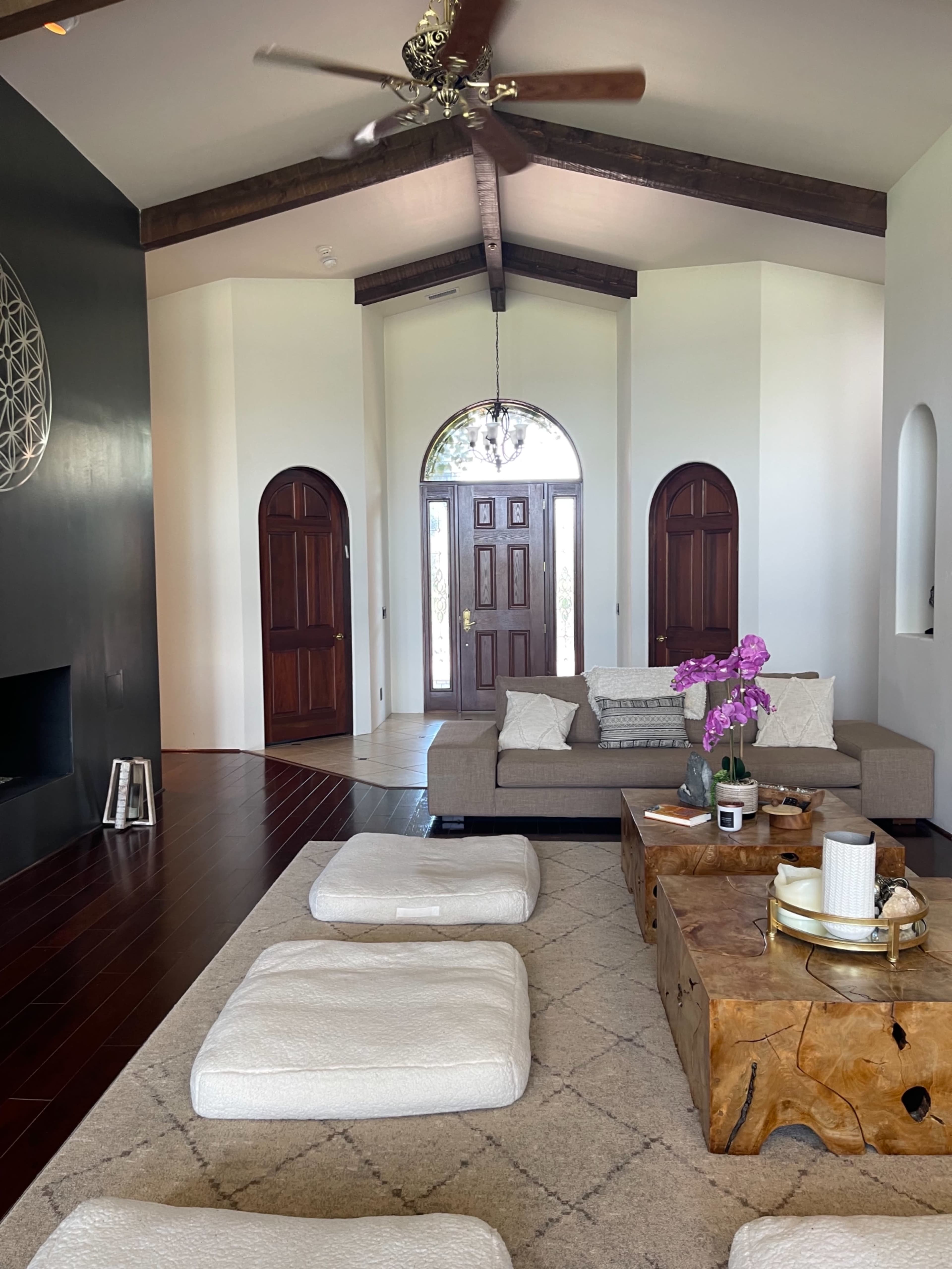 A spacious living room features a large beige sofa, a wooden coffee table, and decorative poufs on a light carpet, with arched doorways leading to the entrance.
