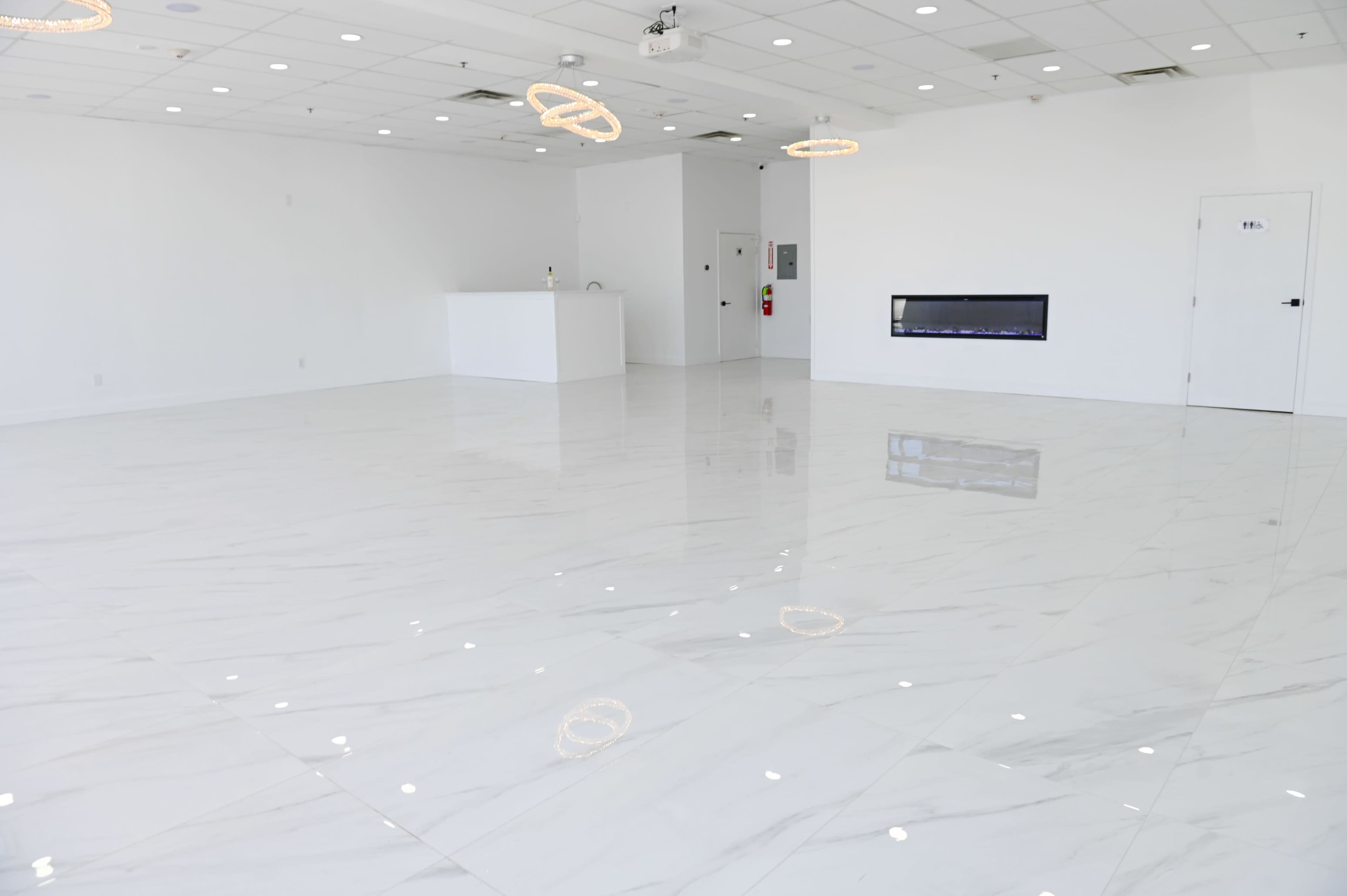 A spacious, empty room with white marble tiles, a recessed fireplace, and two circular light fixtures hanging from the ceiling.
