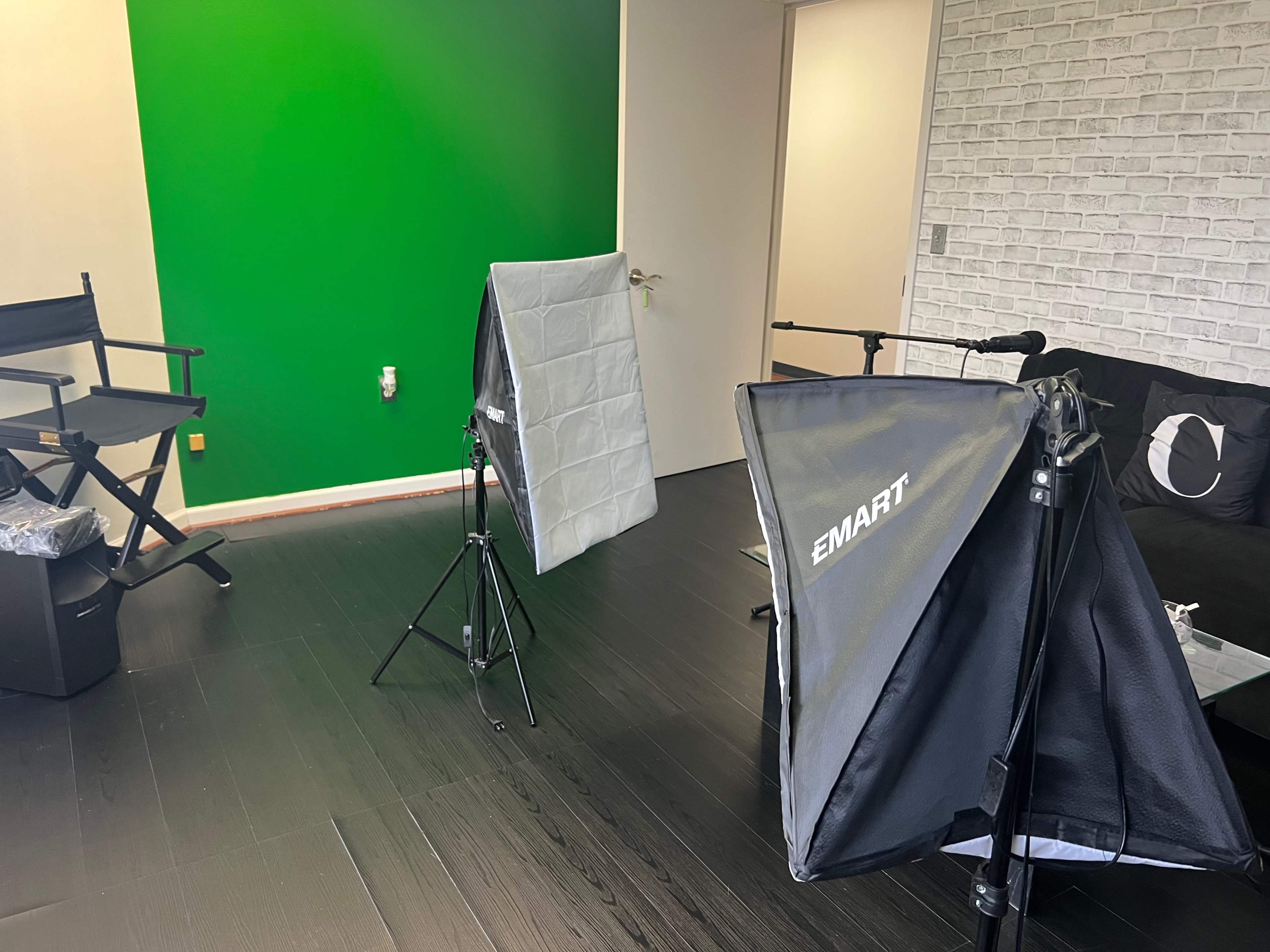 A studio setup with softbox lights, a green screen, and a chair in a room with dark flooring and a white brick wall.