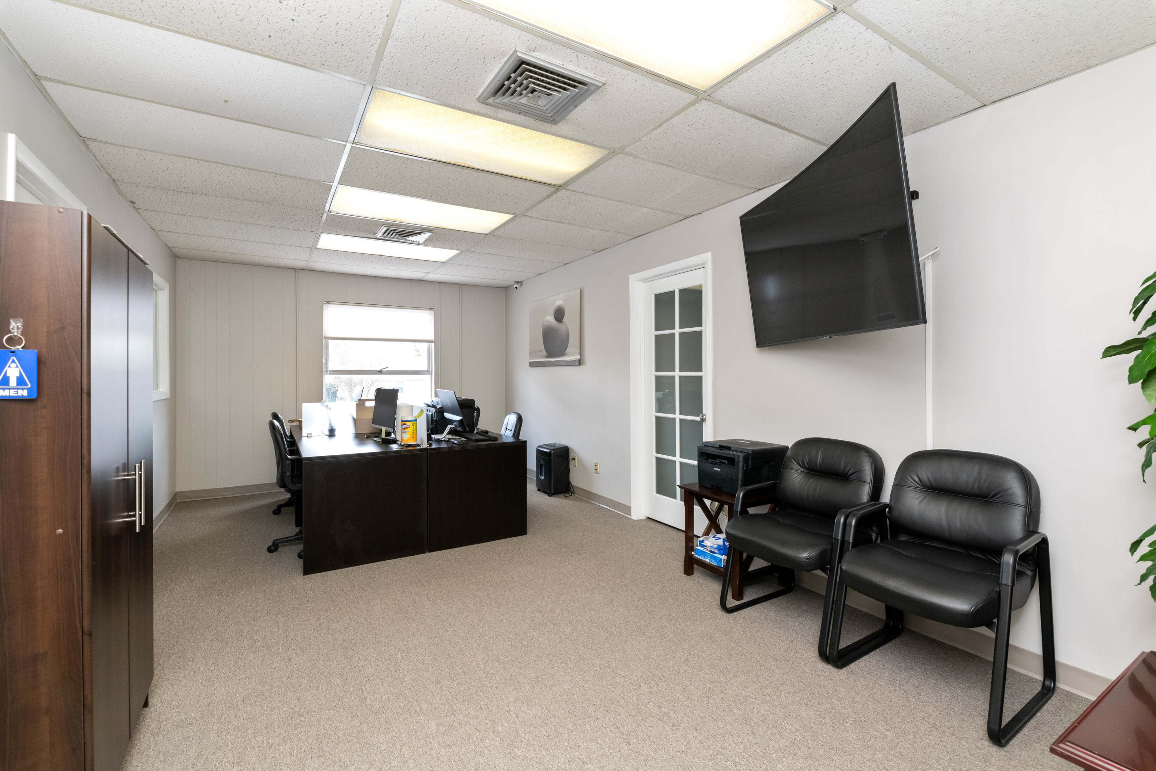 A spacious office with a large desk, an overhead TV, and two black chairs against the wall.